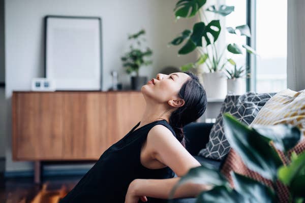 Person relaxing on a couch, leaning back with eyes closed. Indoor plants and modern furniture are in the background, creating a peaceful setting
