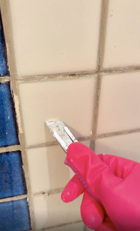 Reddit post showing a gloved hand cleaning tile grout with a razor blade. Viewer questions if it’s soap scum or important part of tile