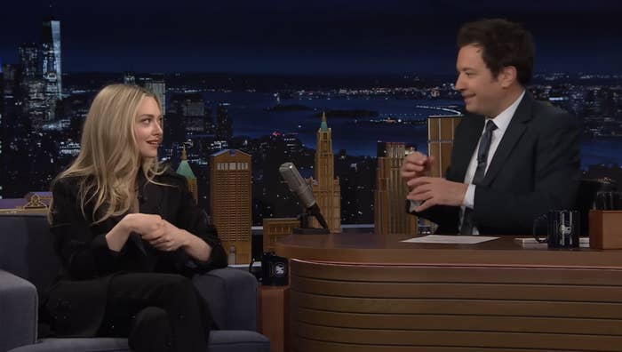 Celebrity guest in a black outfit converses with host at a talk show desk, against a cityscape backdrop