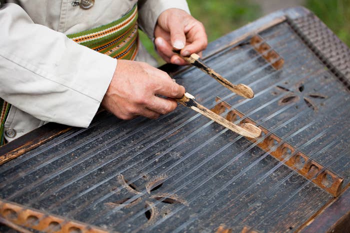 Person playing a traditional string instrument with mallets, wearing a long-sleeved shirt. Intricate patterns are carved on the instrument