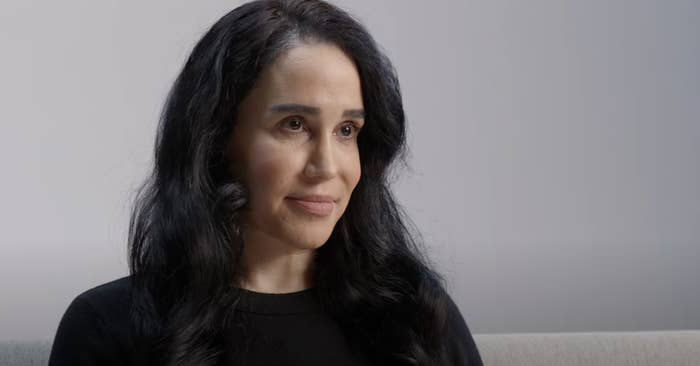 Person with long, dark hair, wearing a black top, sitting indoors, looking slightly to the side