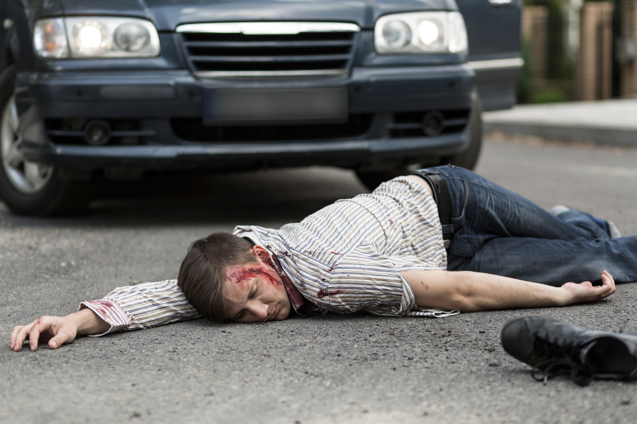 A person lies on a street near a car, appearing injured, with visible blood on their head, suggesting a possible accident