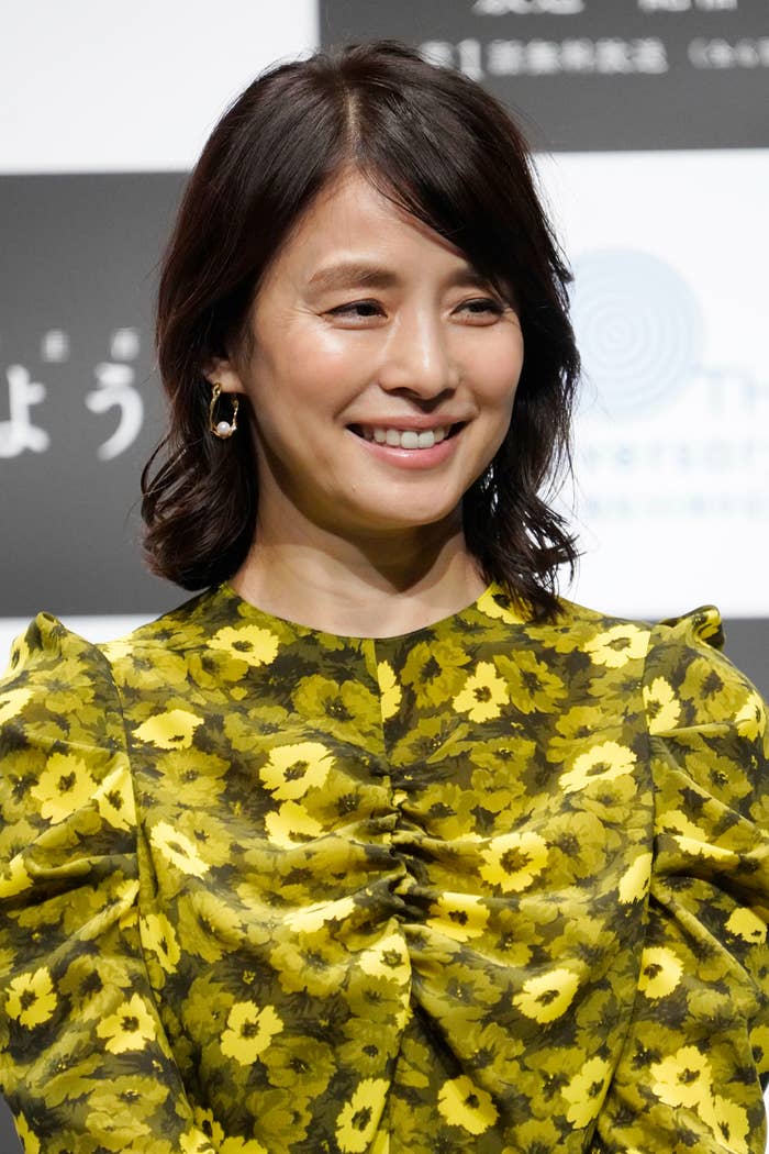 女性が白地に黒の文字がある背景の前で微笑んでいます。彼女は花柄のトップを着ています。