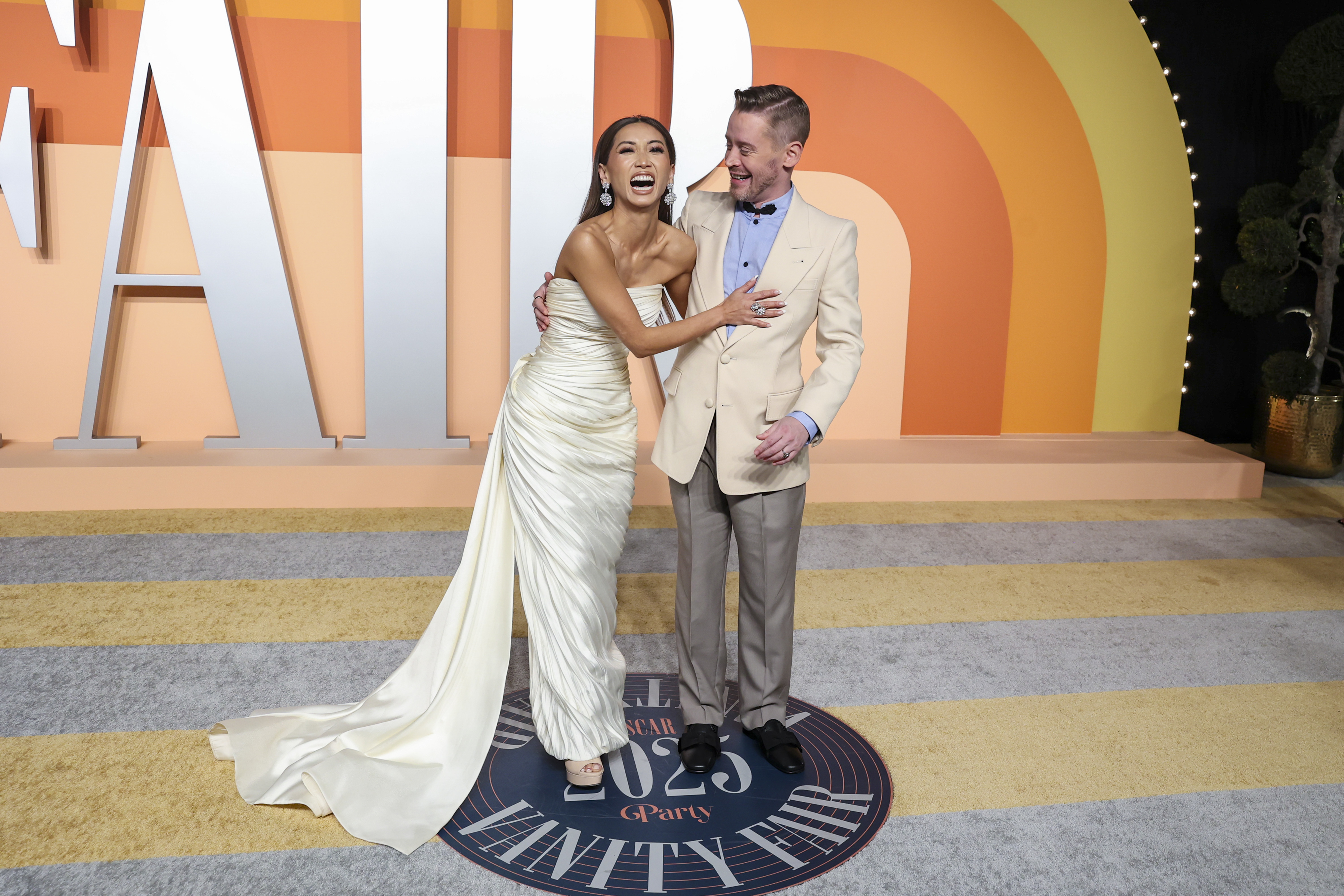 Two people on a red carpet, smiling and interacting. One wears an elegant strapless gown; the other wears a light suit with a blue shirt. Vanity Fair event