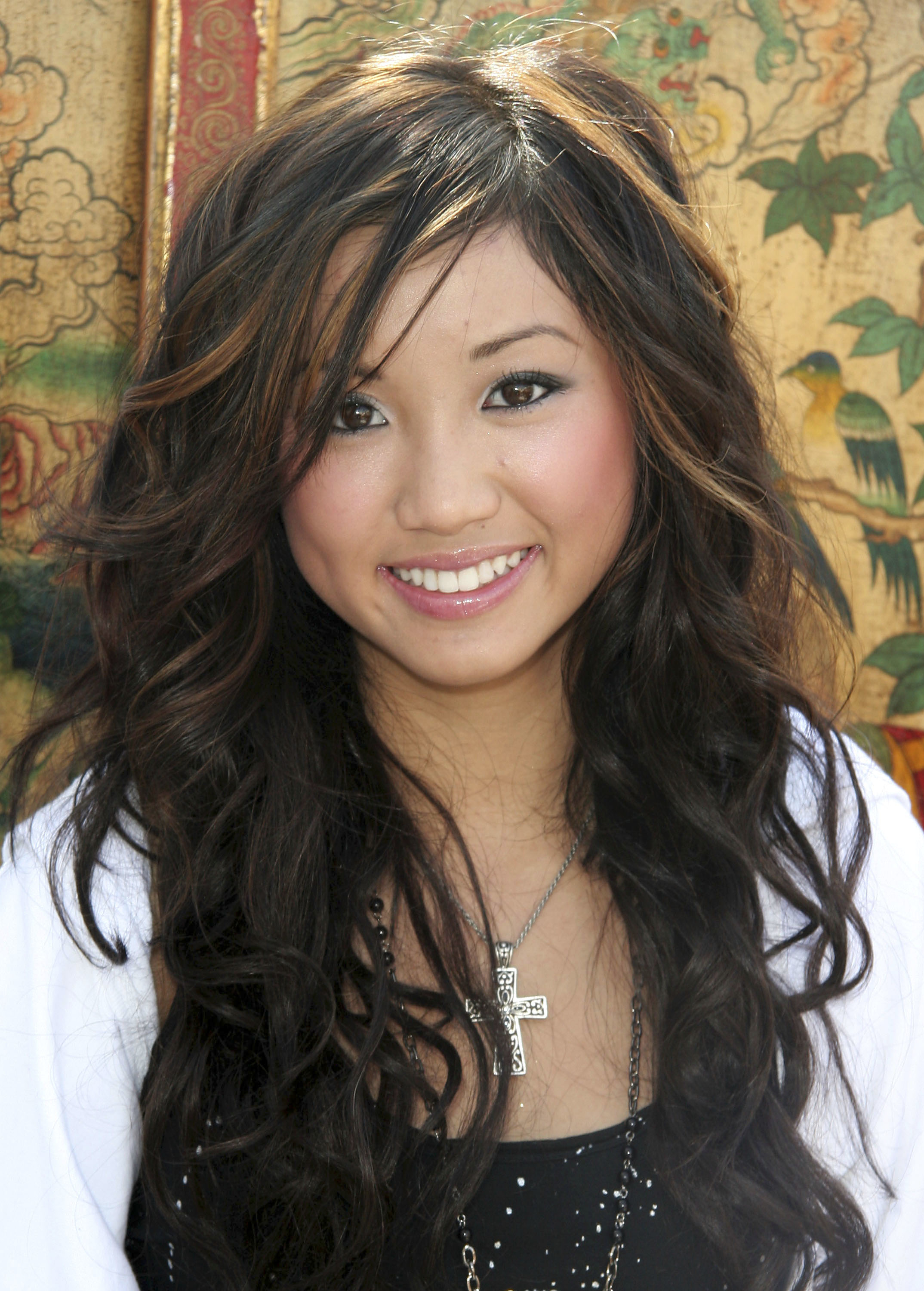 Person with long, wavy hair wearing a cross necklace and a light wrap, standing in front of a decorative background