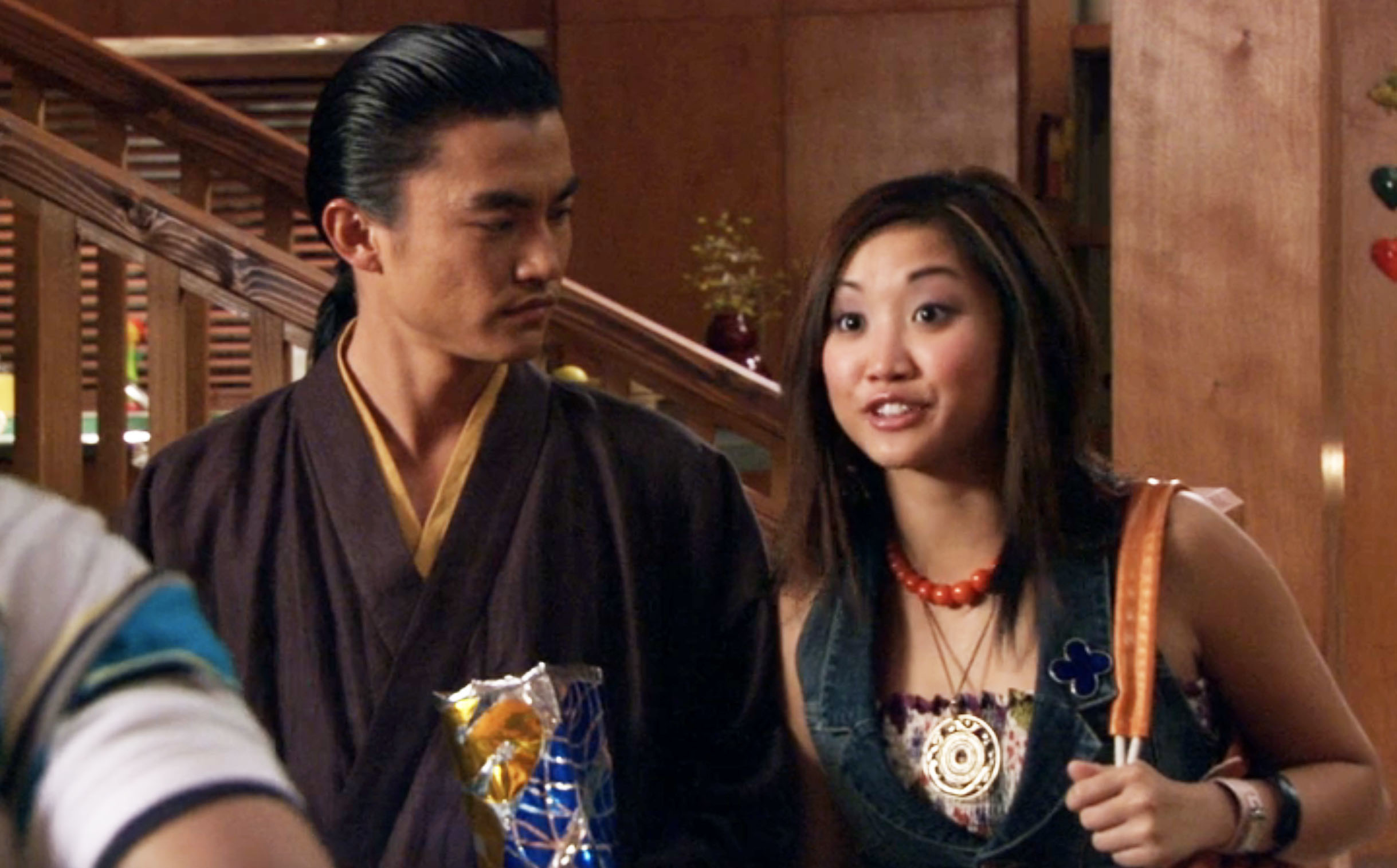 Two people in casual attire, one holding snacks, stand in a home setting, possibly in a scene from a TV show or movie