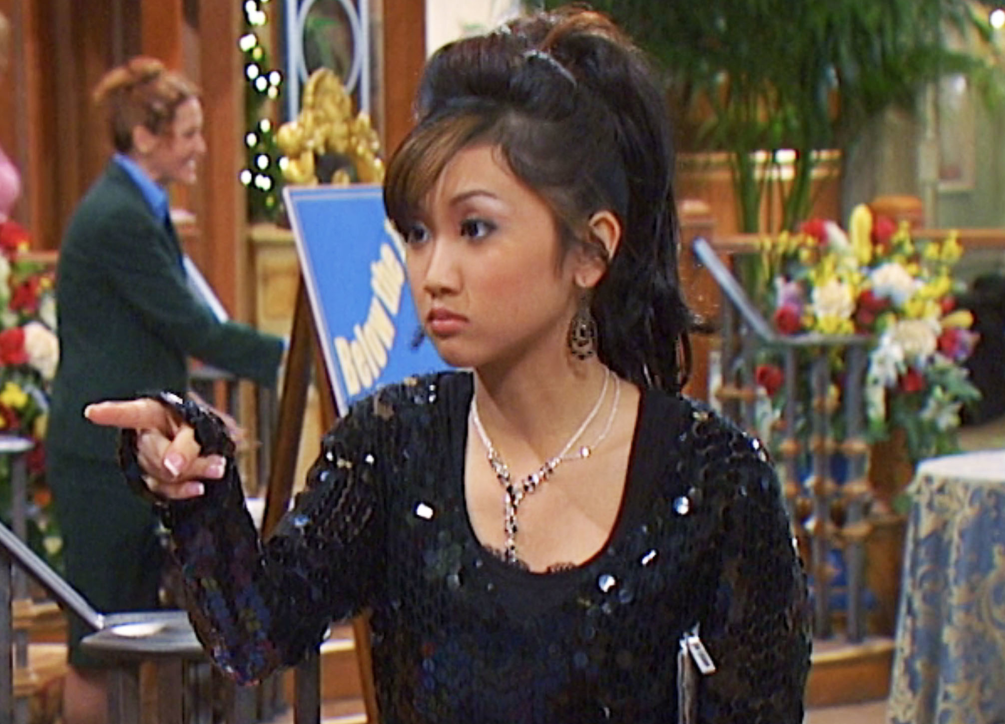 Person in a sparkly outfit pointing, in a setting resembling a hotel lobby, with flowers and a staircase in the background