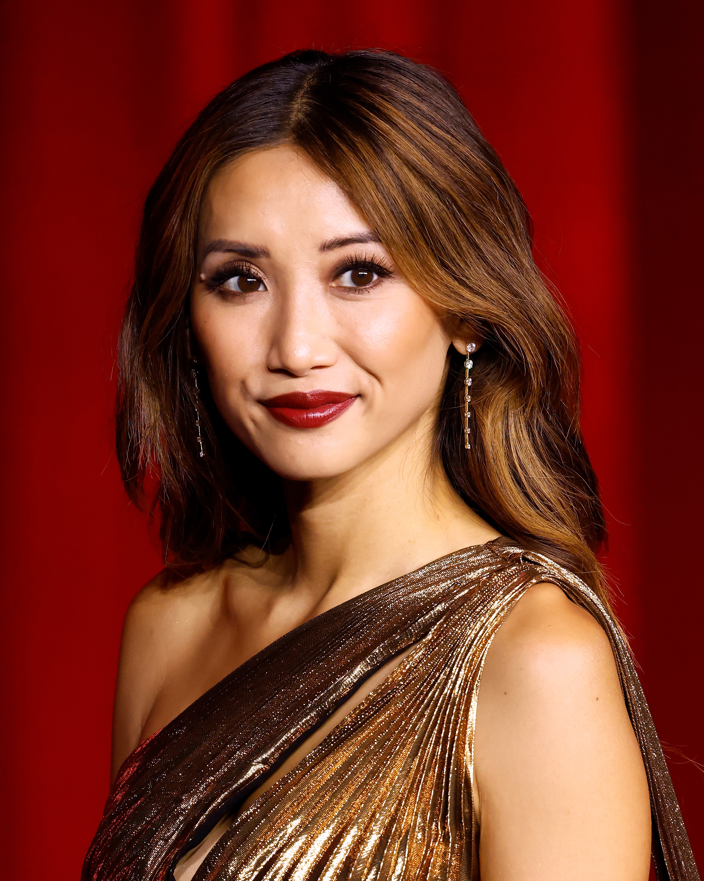 Person in elegant, shimmering gown with asymmetrical neckline, standing against a red backdrop at an event