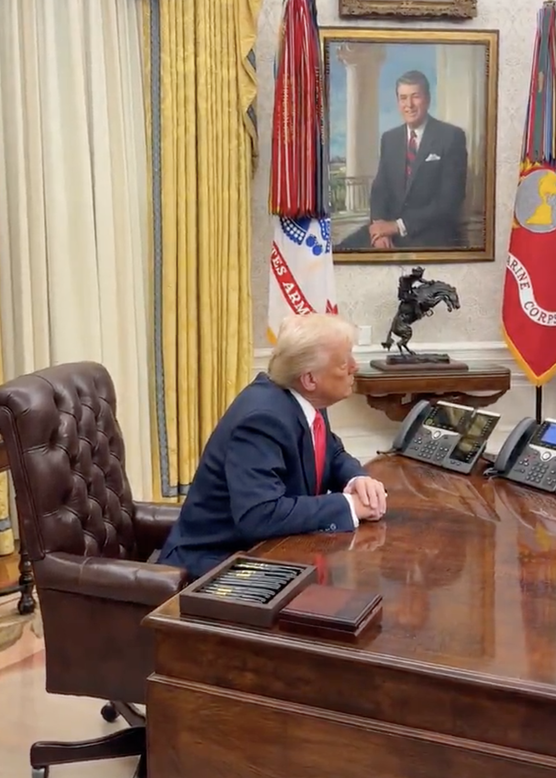 Person seated at a large wooden desk in an office with government flags and a portrait in the background, speaking on the phone