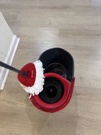 A mop with a reddish grip and mop caput is placed successful a reddish and achromatic bucket connected a woody floor, suitable for household cleaning