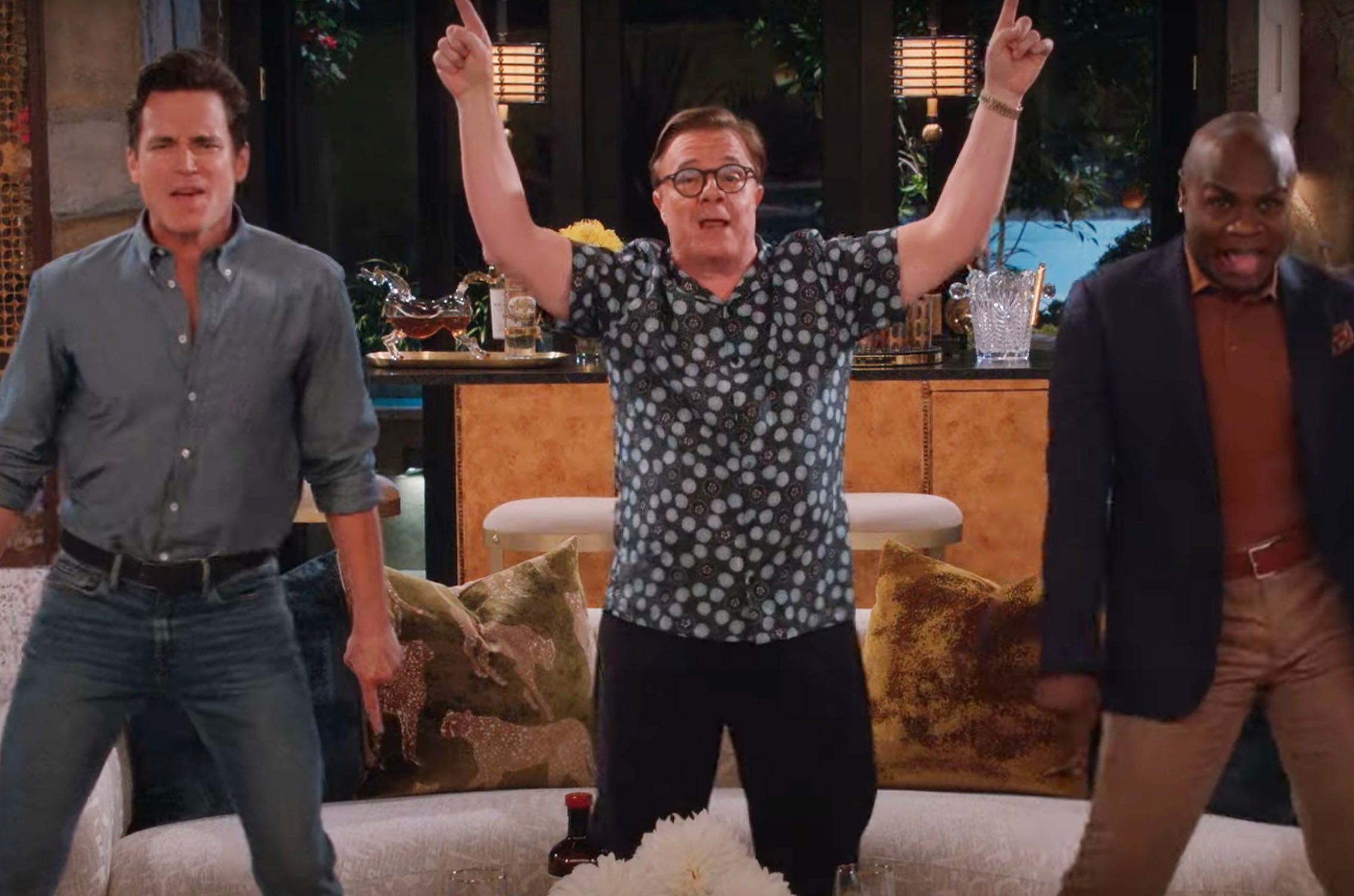 Three people in a living room set, performing enthusiastically, with one raising arms. Casual and relaxed attire is worn by all