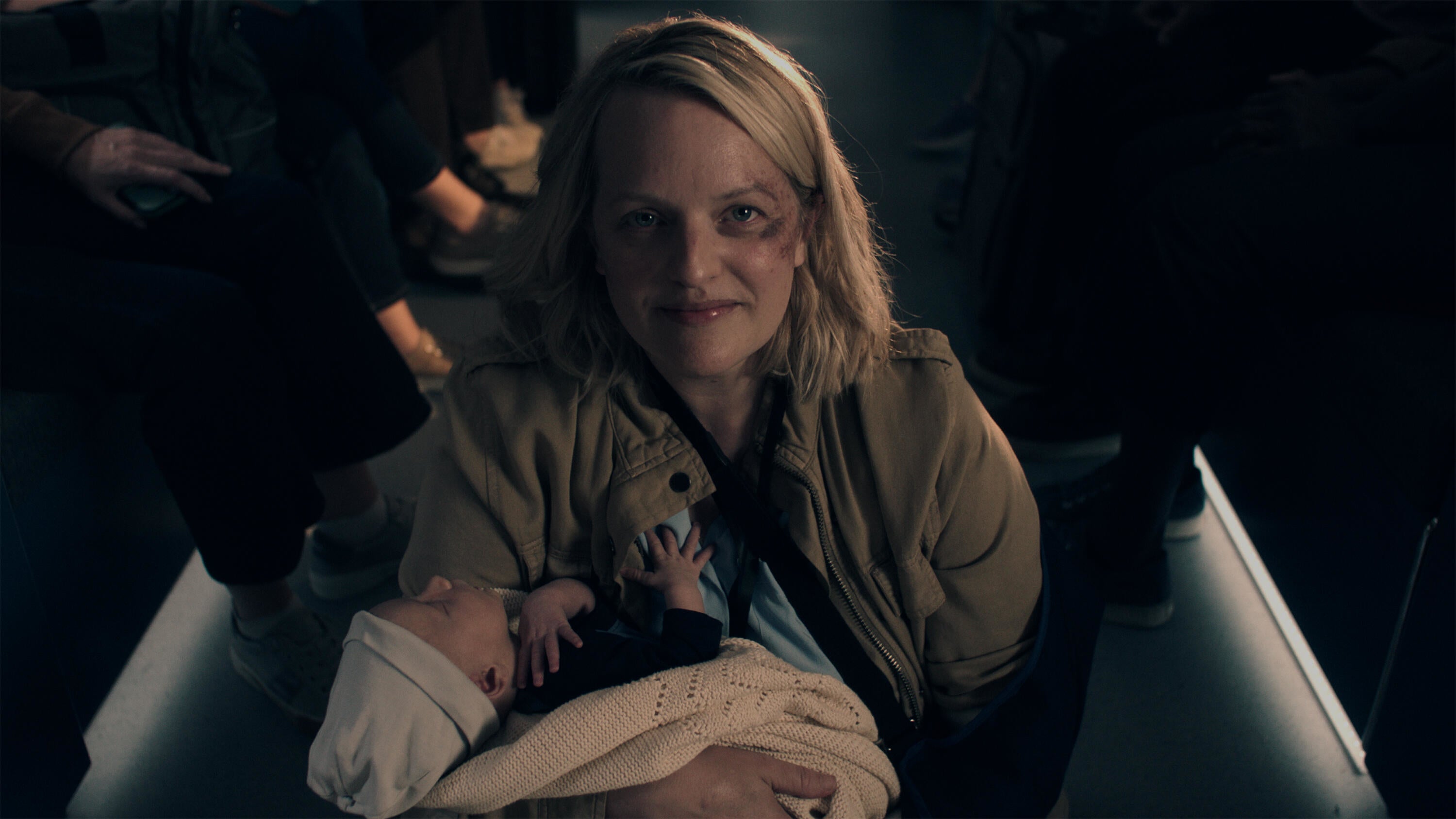 Elisabeth Moss as June holding a baby, sitting in a dimly lit environment, smiling gently at the camera