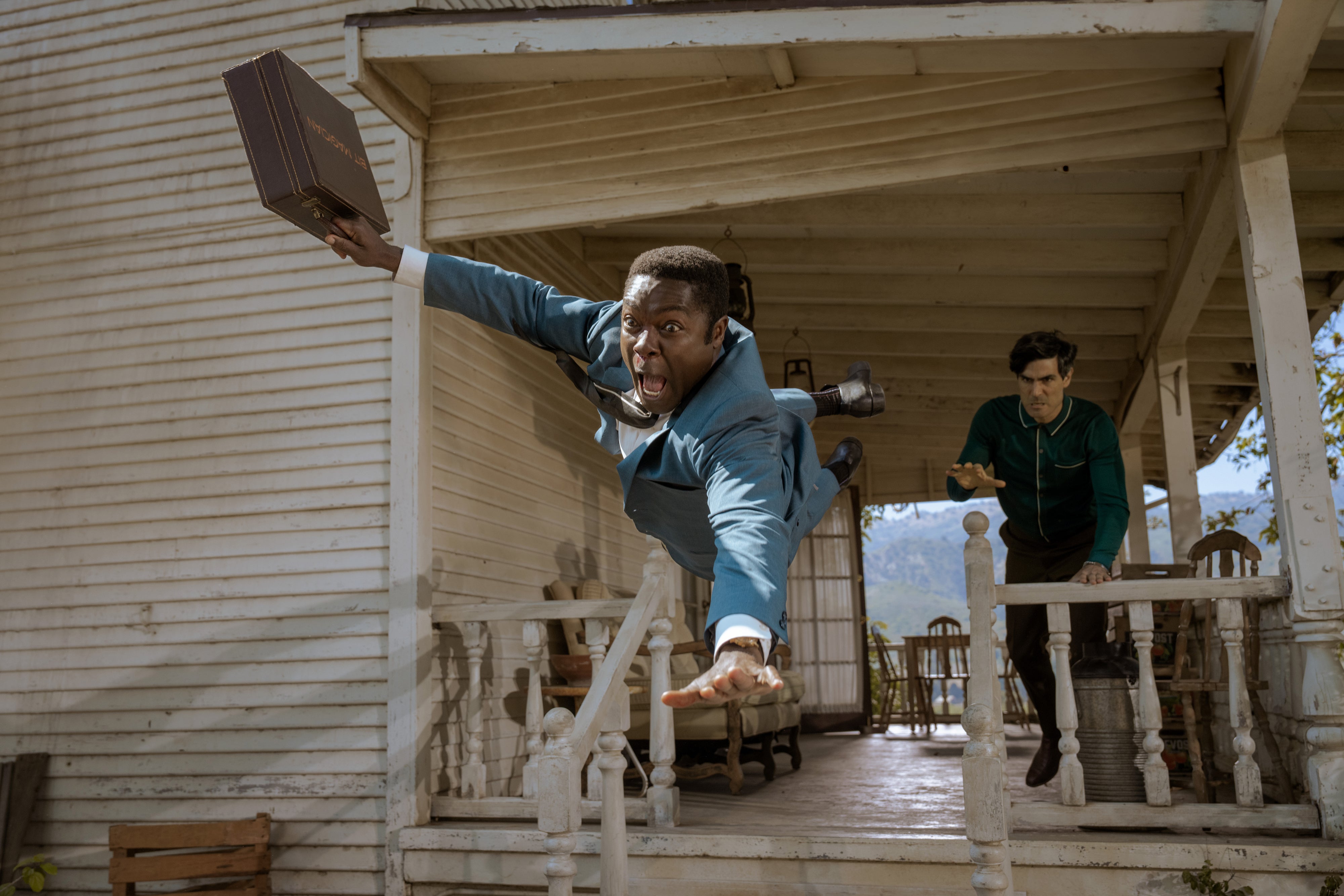 Man in a suit dramatically leaps off a porch holding a briefcase, with another person in the background looking surprised in a scene from "Government Cheese"