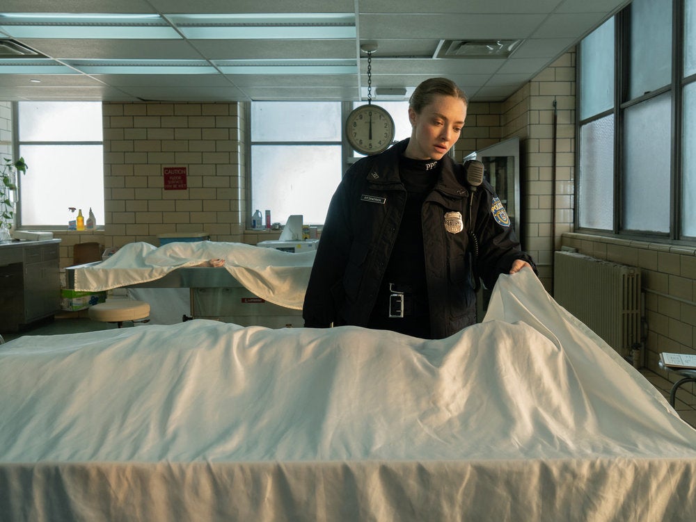 Amanda Seyfried as a police officer stands in a morgue, lifting a sheet from a body on a table
