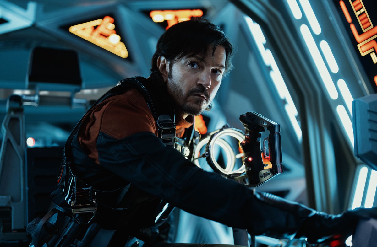 Diego Luna as Cassian Andor in a futuristic spaceship cockpit, wearing a black and red suit, focused on operating controls