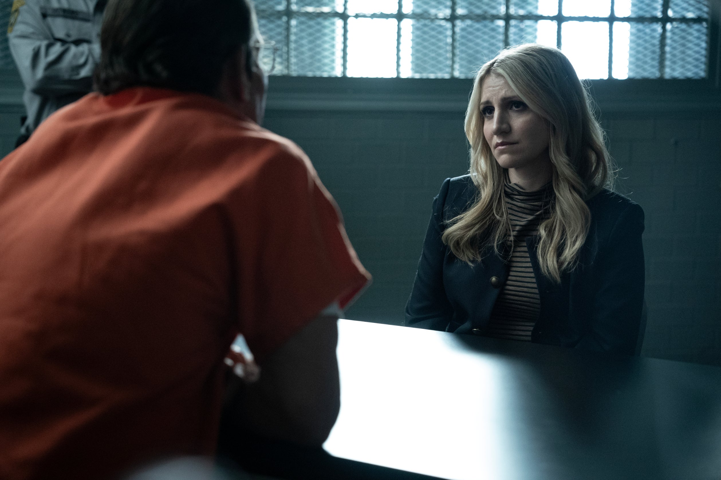 A woman in a blazer sits across a man in an orange jumpsuit, in a prison visiting room