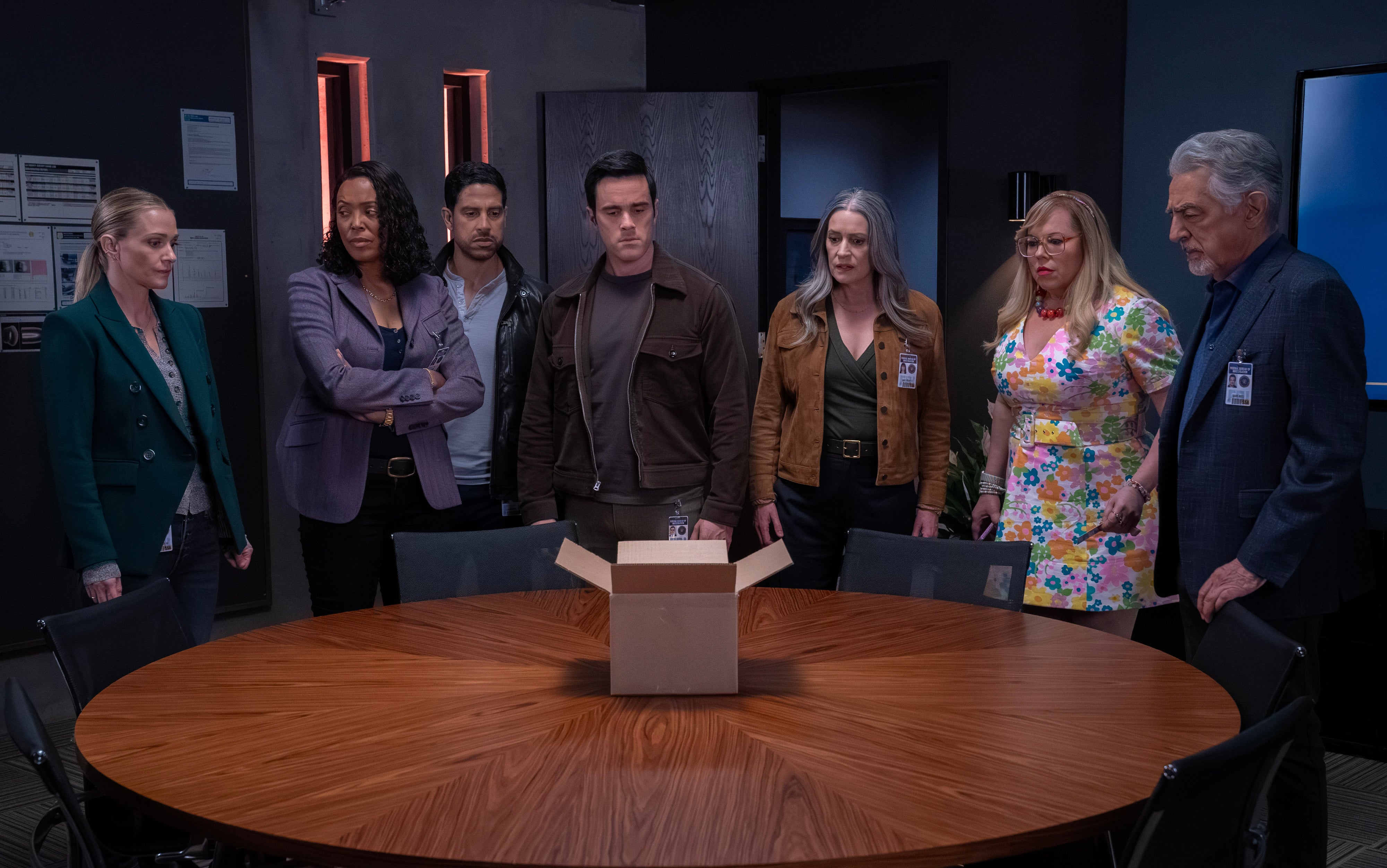 Group of seven people in a meeting room intently looking at an open box on a round table, suggesting a discussion or investigation scene