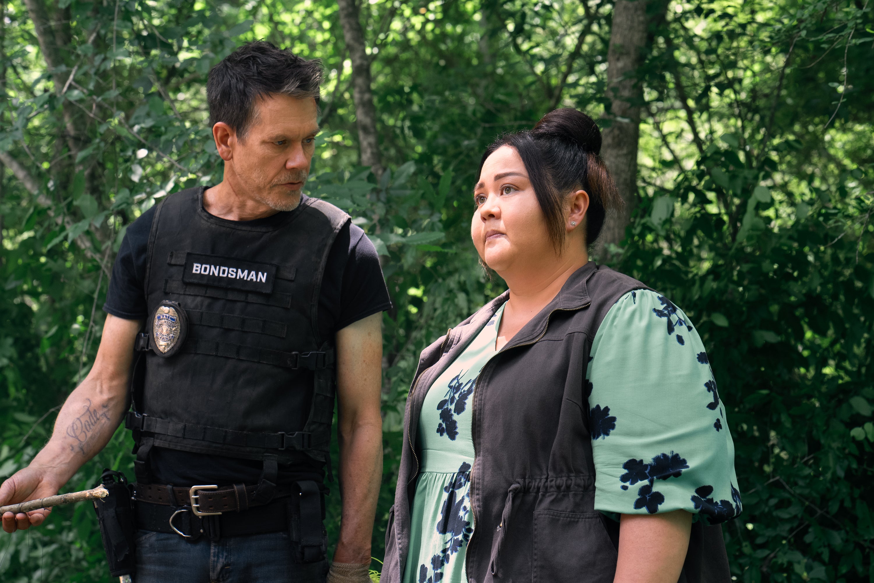 Two actors in a forest scene; one wears a tactical vest labeled "Bondsman," and the other wears a floral dress with a vest