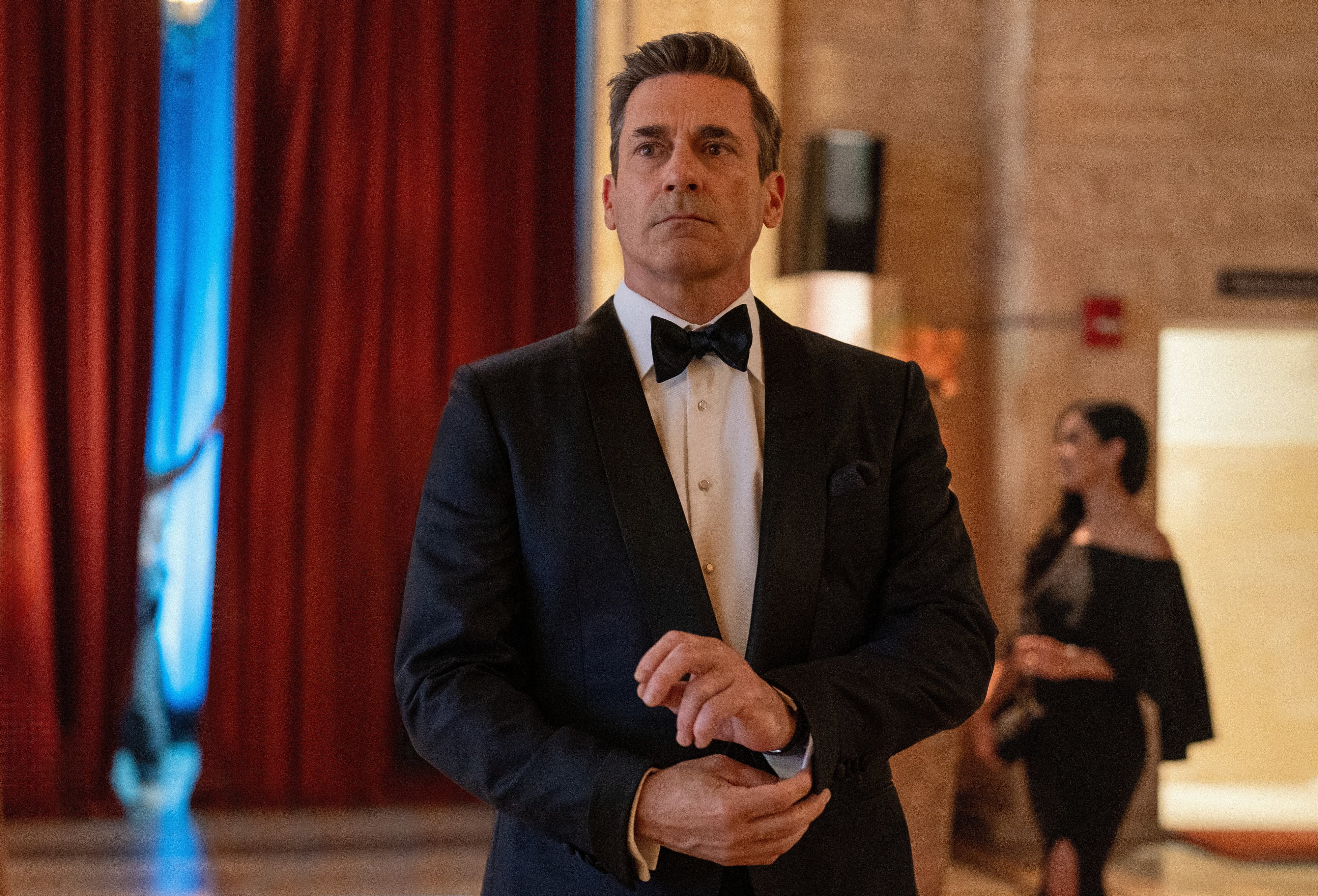 Jon Hamm in a black tuxedo adjusts his cufflink at an elegant event in a scene from a show