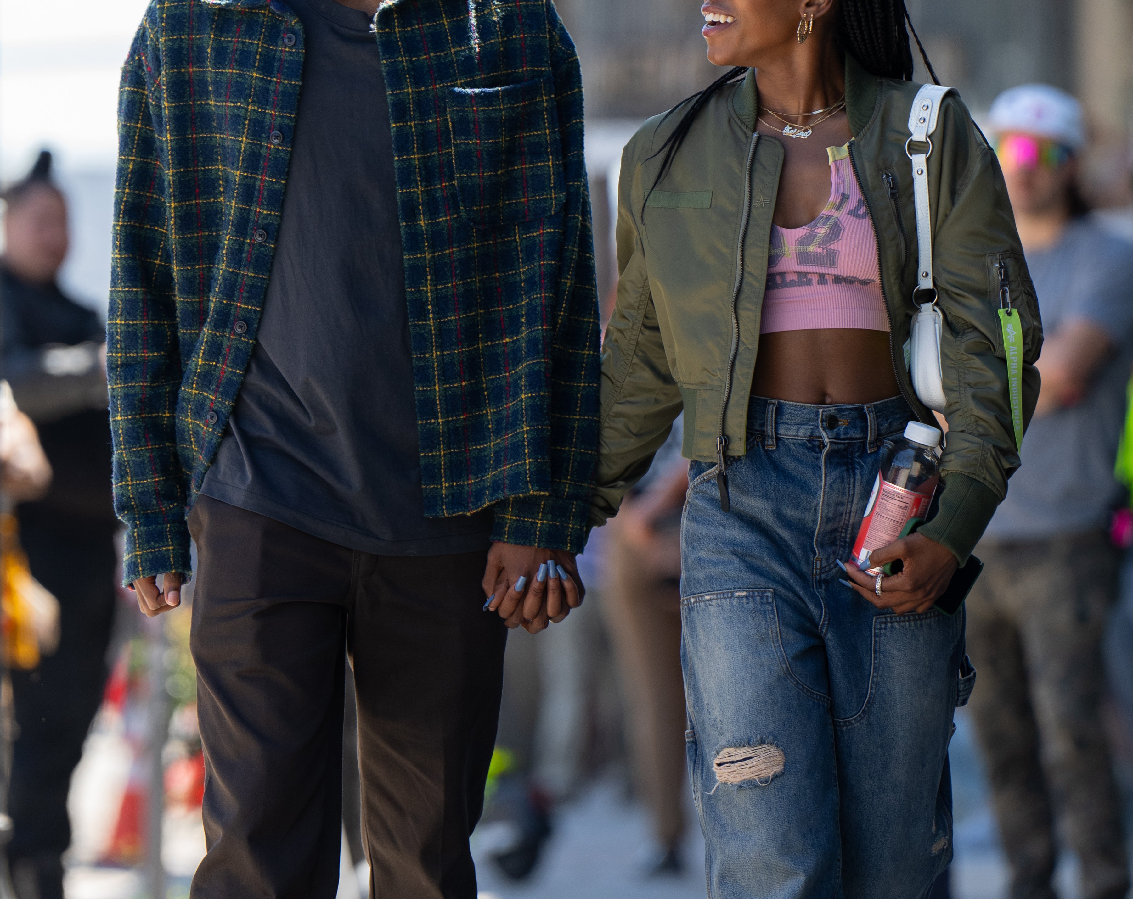 Two people walking hand in hand, smiling at each other. One wears a plaid jacket, and the other wears a green jacket and wide-leg jeans