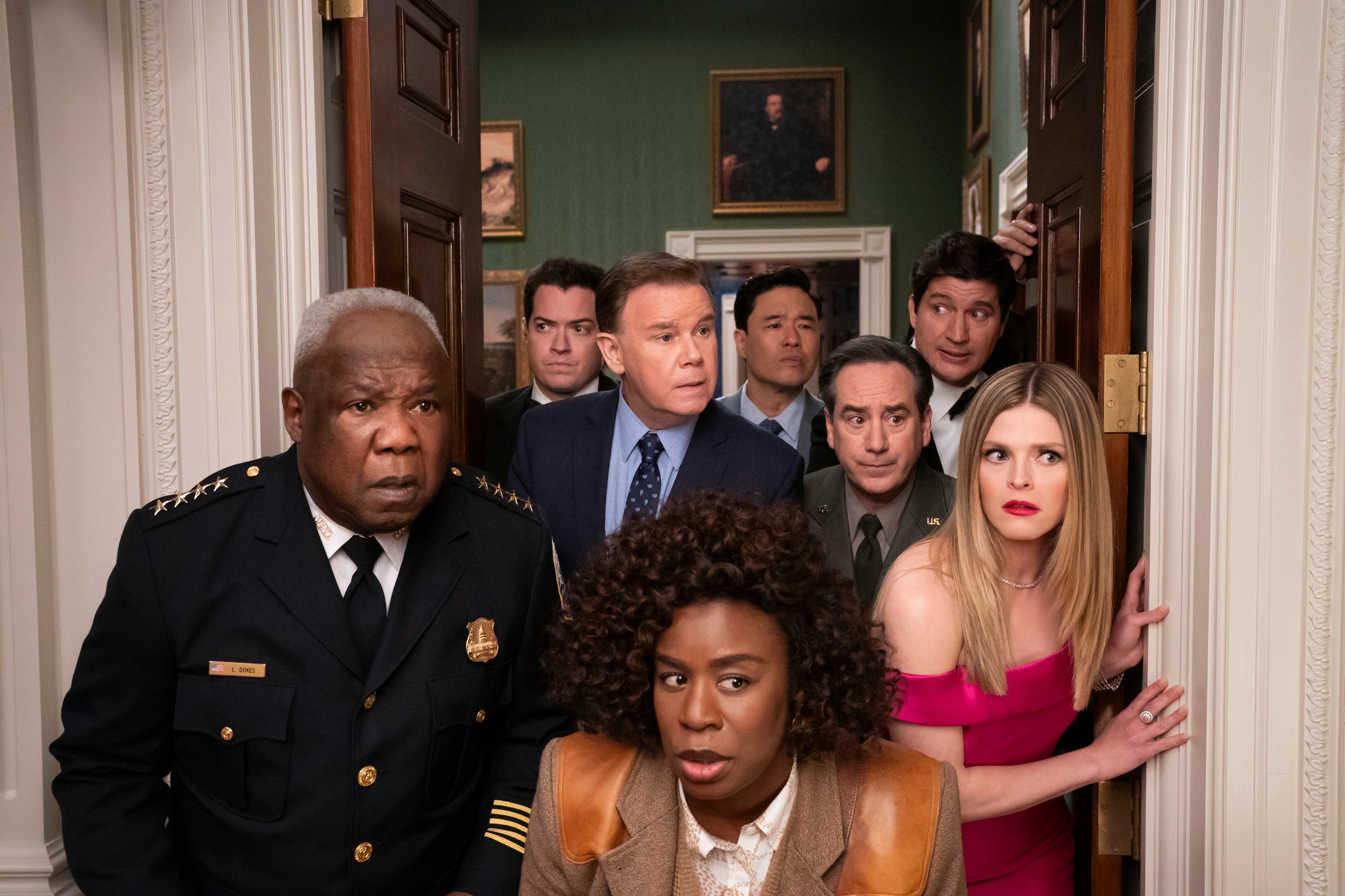 A group of people in suits and formal wear gather in a hallway, looking surprised. They appear to be listening into a conversation
