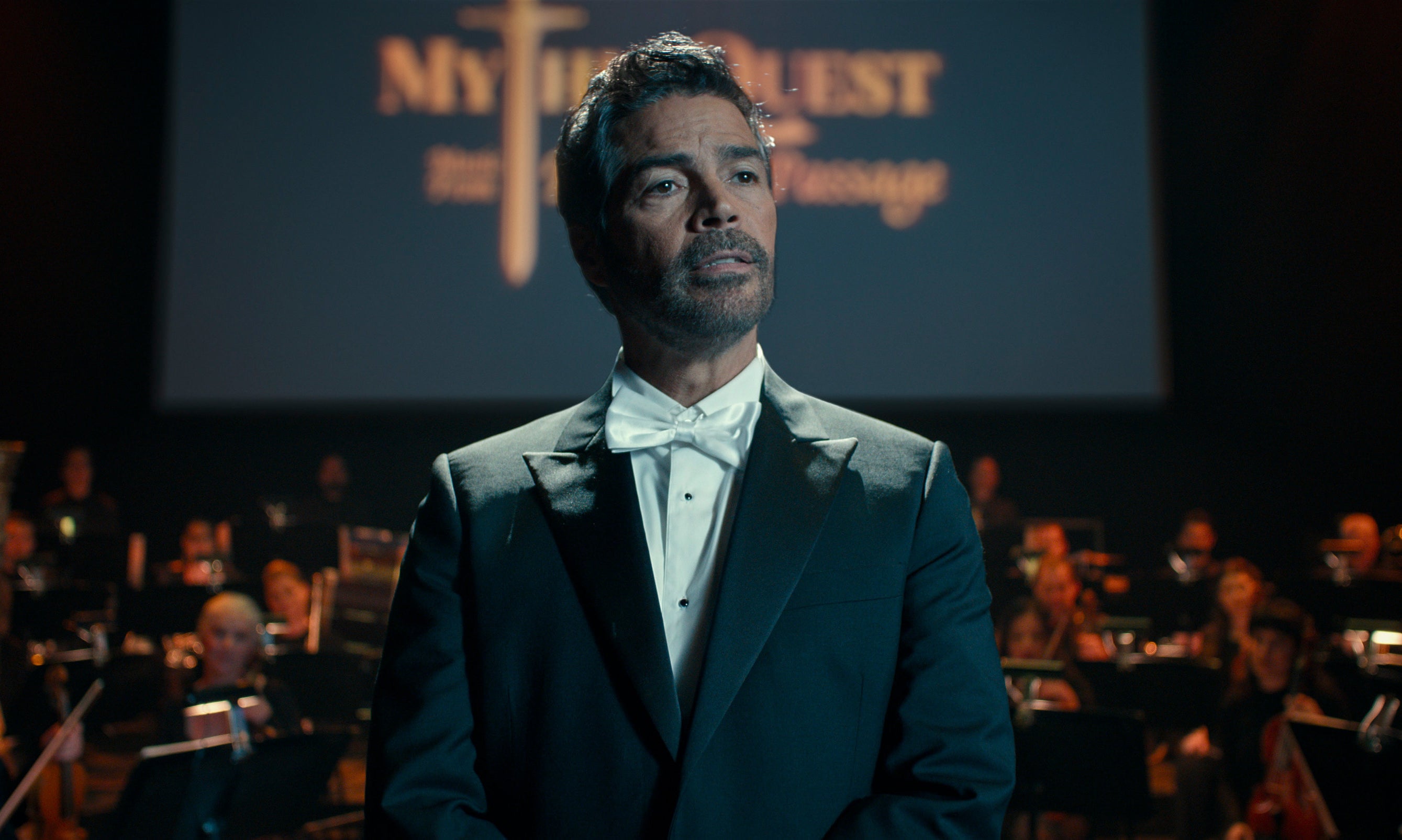 A man in a tuxedo stands in front of an orchestra with a screen displaying "Mythic Quest" behind him