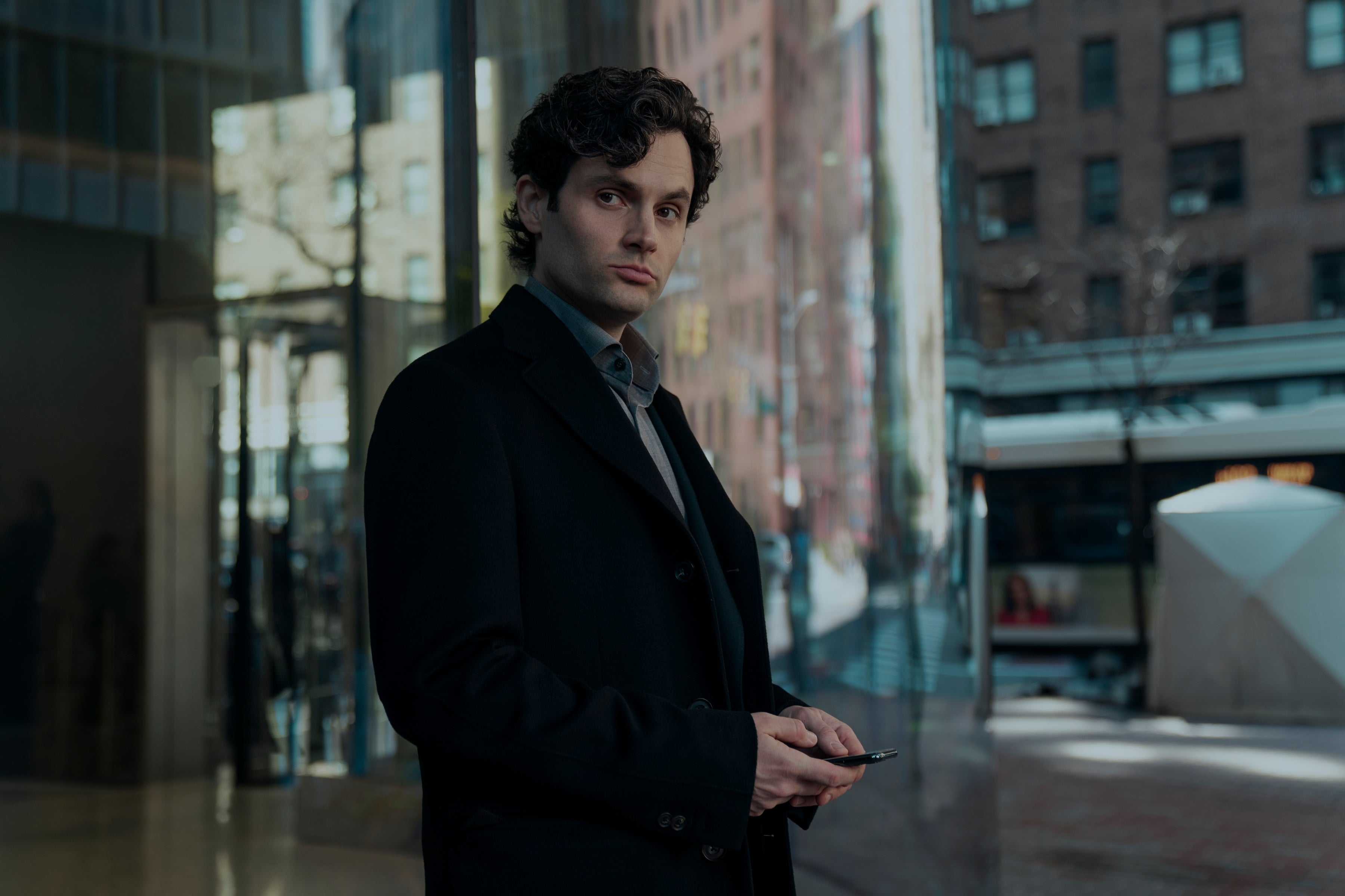 Penn Badgley as Joe Goldberg standing on a street corner in NYC holding his phone and looking suspicious