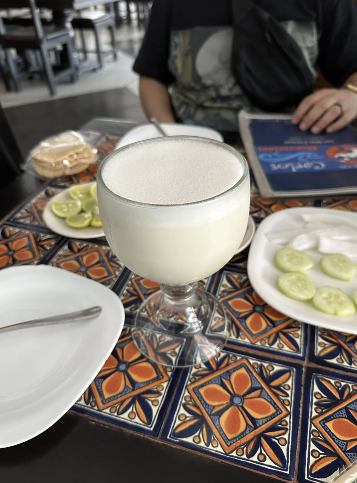 Et bord med en pina colada i et glass, pyntet med kokosnøttflak. Bakgrunn viser en persons overkropp og en annen rett med kalkskiver