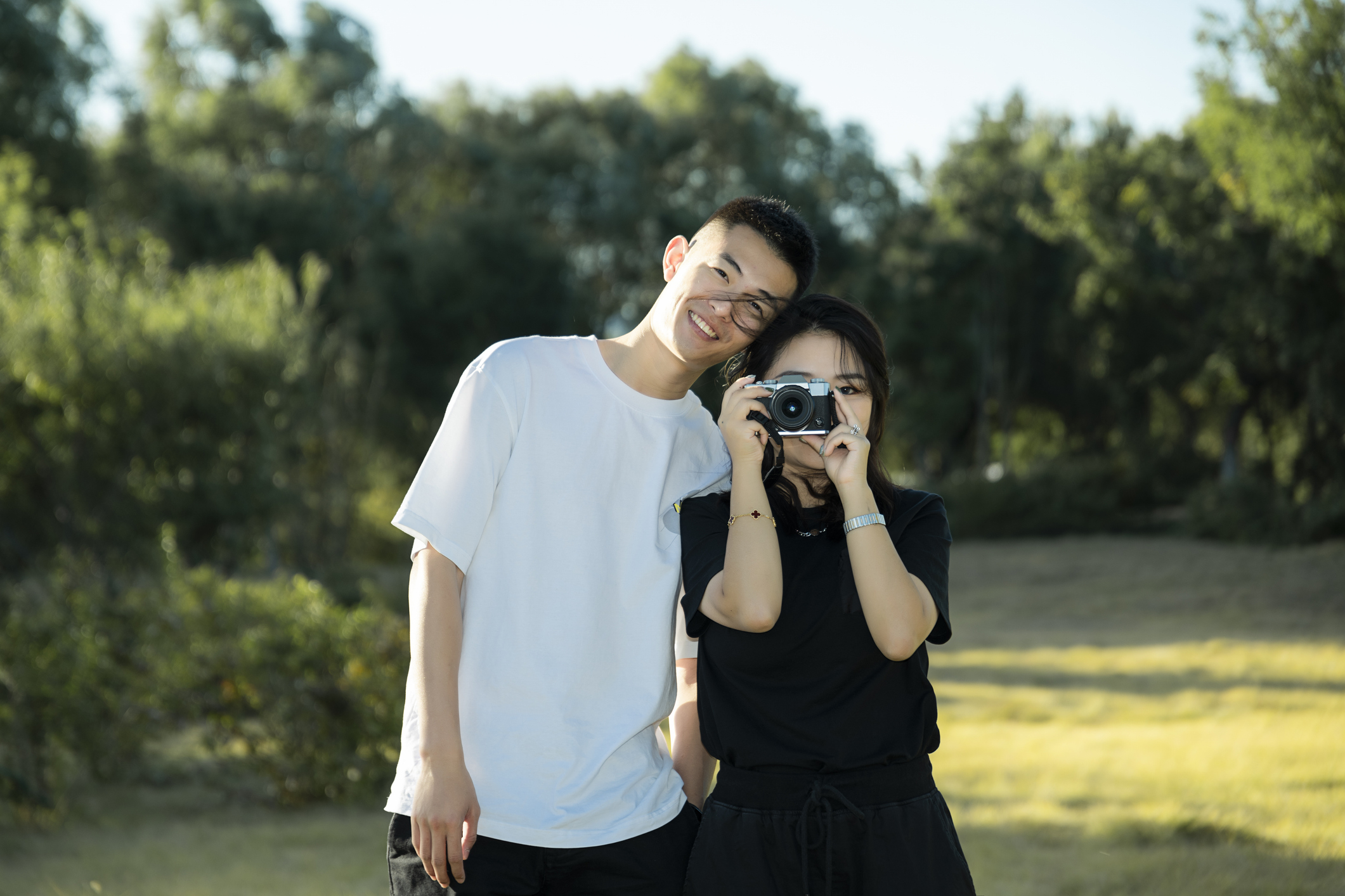 En smilende person i en hvit skjorte lener seg mot en person i en svart skjorte som holder et kamera utendørs