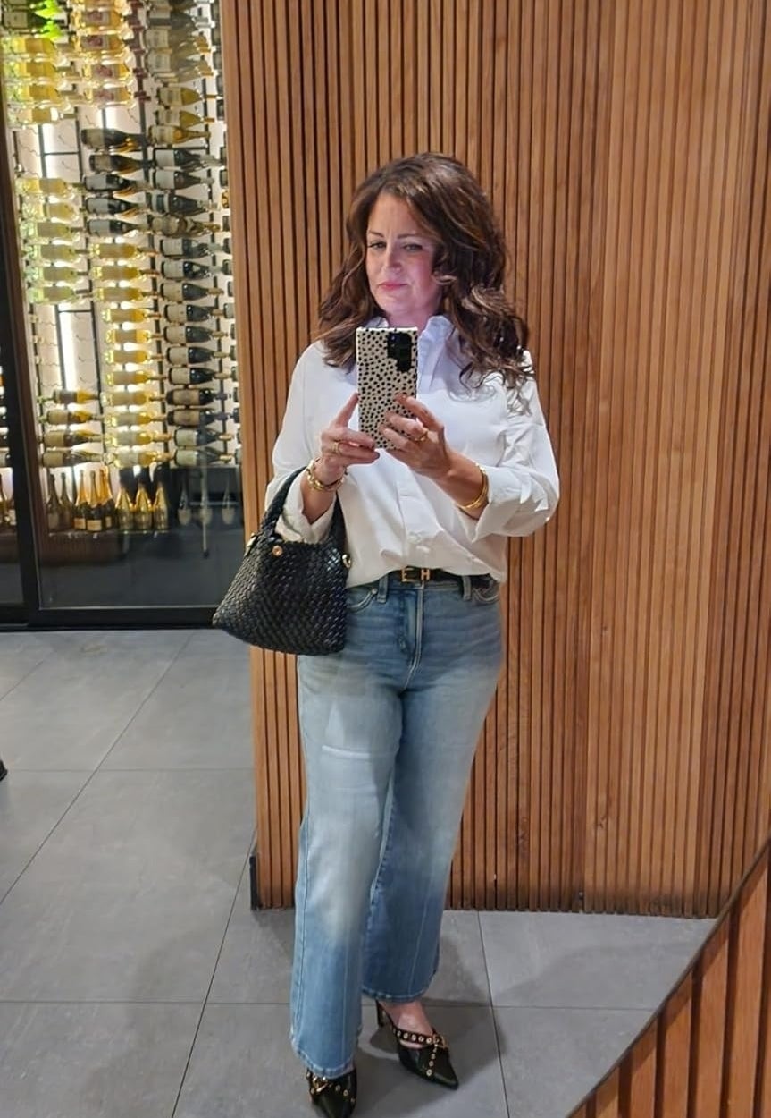 Wanita dengan blus putih dan jeans memegang telepon, merefleksikan di cermin berpanel kayu. Botol anggur ditampilkan di latar belakang