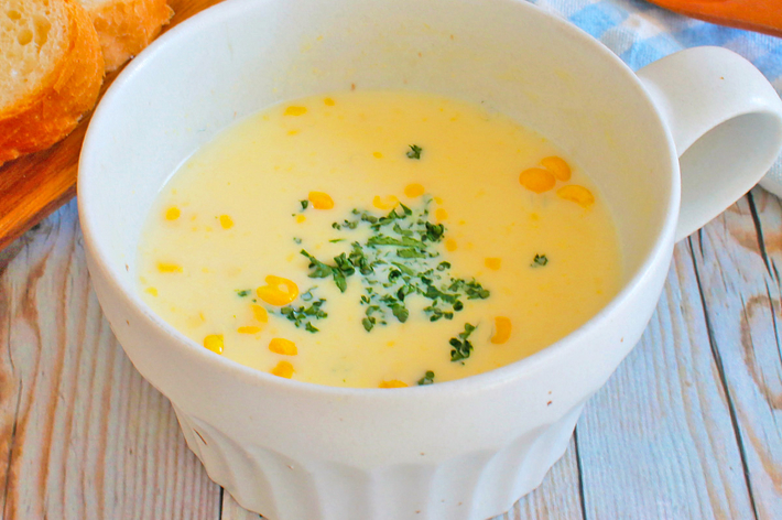 カップに入ったコーンポタージュスープ。上にパセリが散らされ、横にパンとスプーンが置かれているテーブルセッティング。