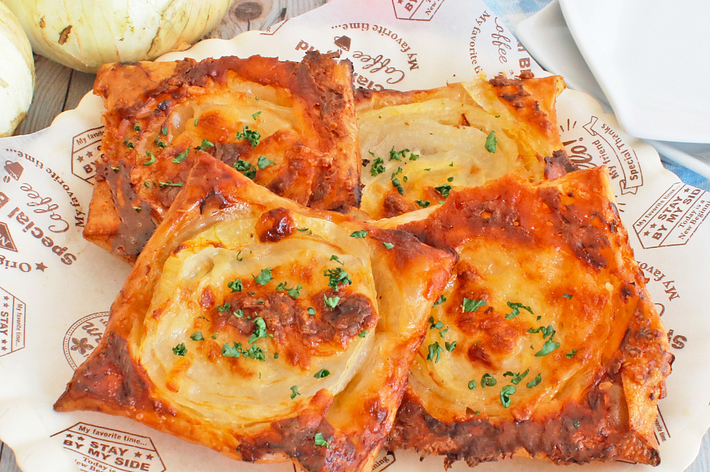 玉ねぎとチーズをトッピングされた四角いパイが4つ、紙の上に置かれている。背景に玉ねぎと皿がある。