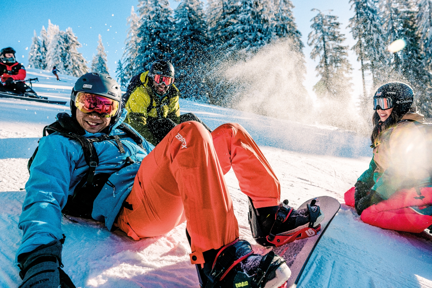 People in ski gear enjoying a snowy mountain. One person sits in the foreground, smiling, while others ski or relax in the background