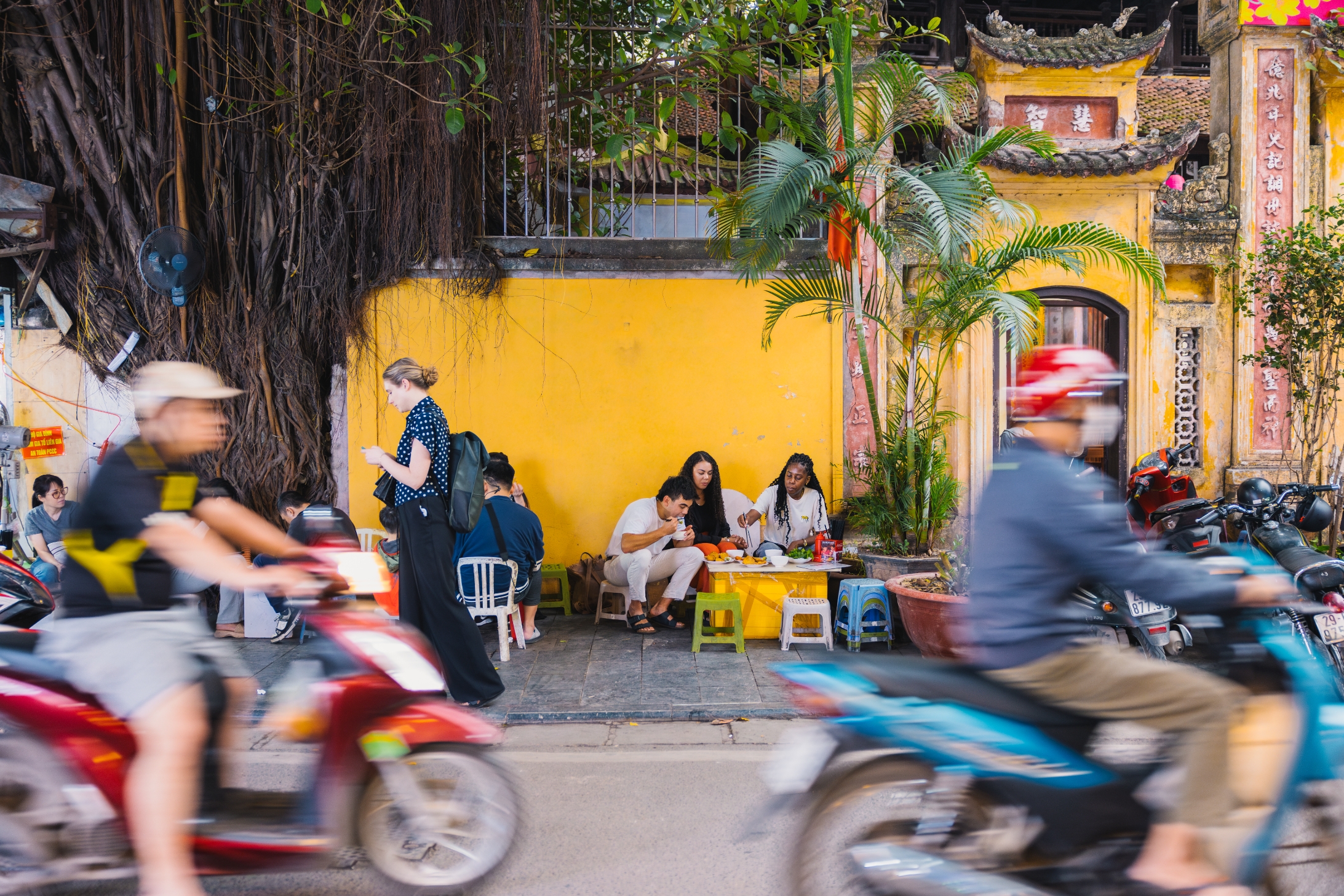 Street scene with people dining at a small outdoor café. Motorcyclists ride by, creating a dynamic contrast with the relaxed atmosphere