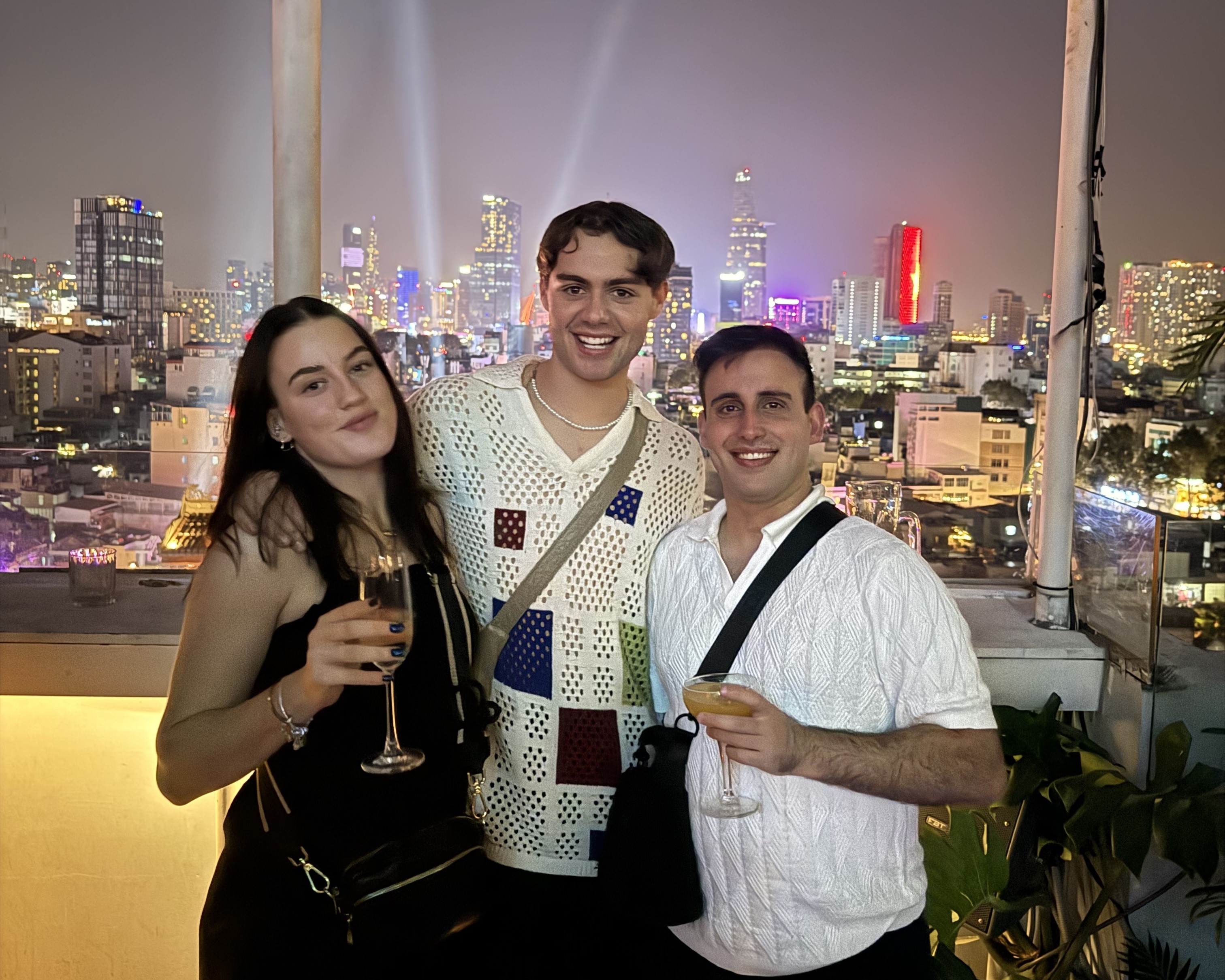 Three people smiling and holding drinks on a rooftop at night with a city skyline and lights in the background