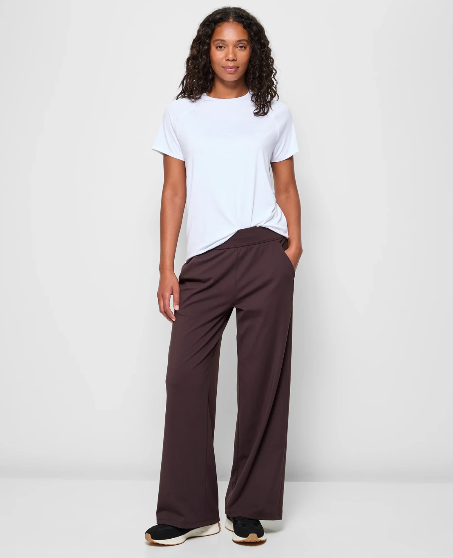 Person in a casual outfit with a white t-shirt, brown wide-leg pants, and sneakers, standing with one hand in their pocket, smiling slightly