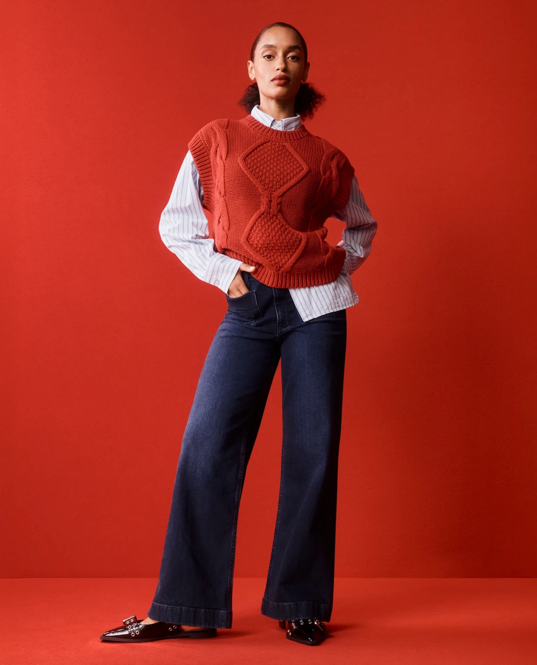 Person posing against a backdrop, wearing a textured sweater over a striped blouse and wide-leg pants