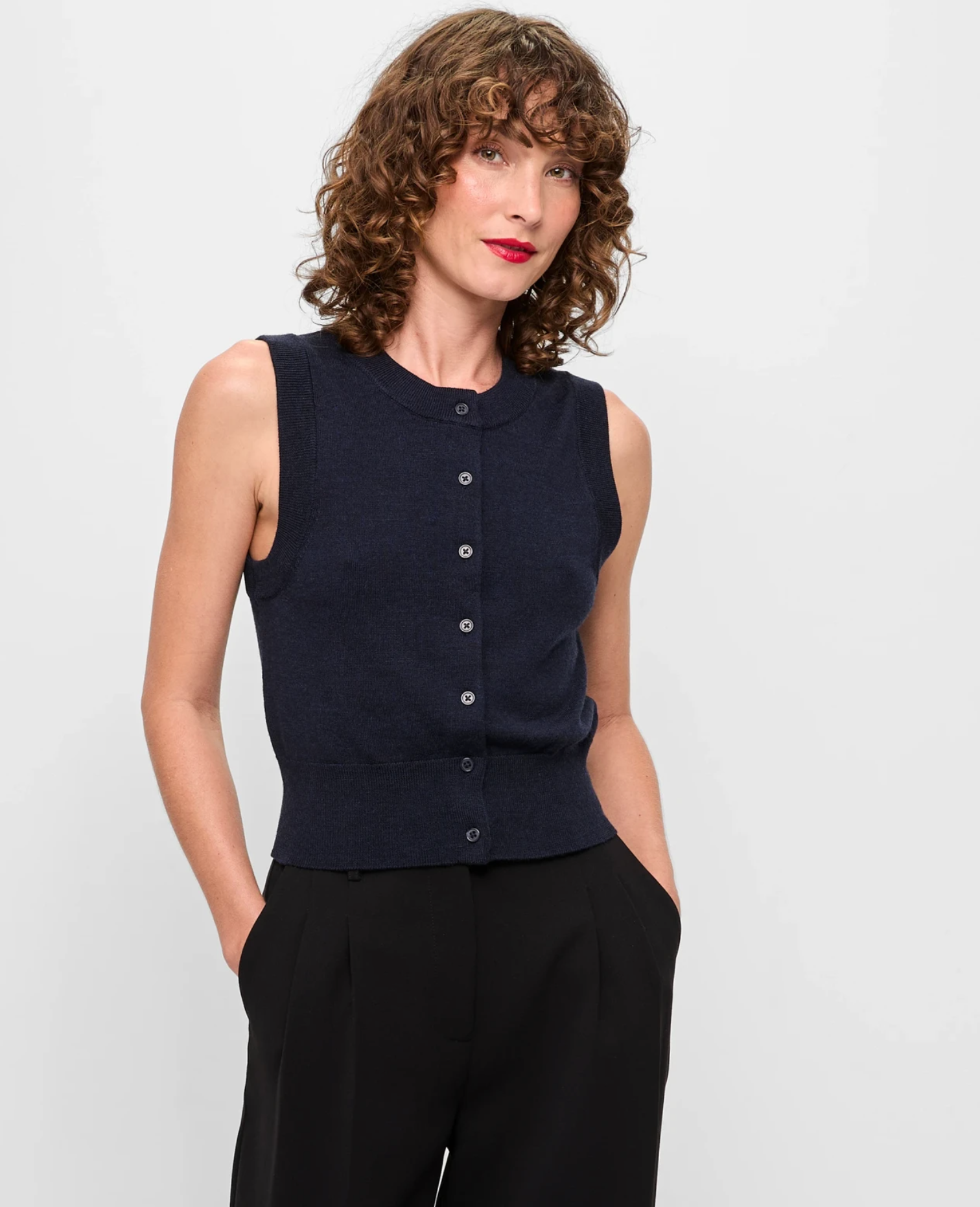 Person posing with hands in pockets, wearing a button-up sleeveless top and pants, styled with curly hair