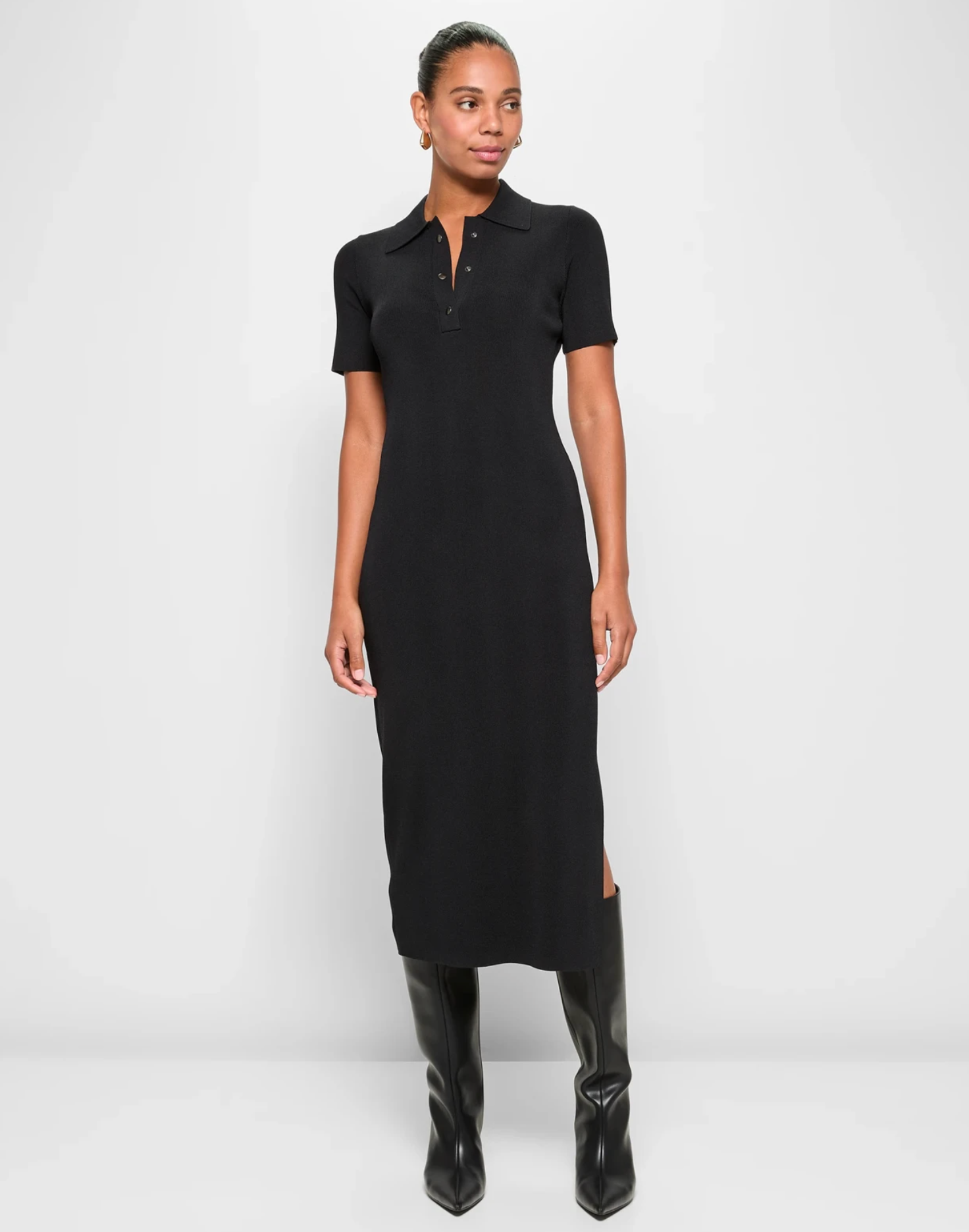 Person in a short-sleeve, button-up, black midi dress paired with knee-high boots, standing against a plain backdrop