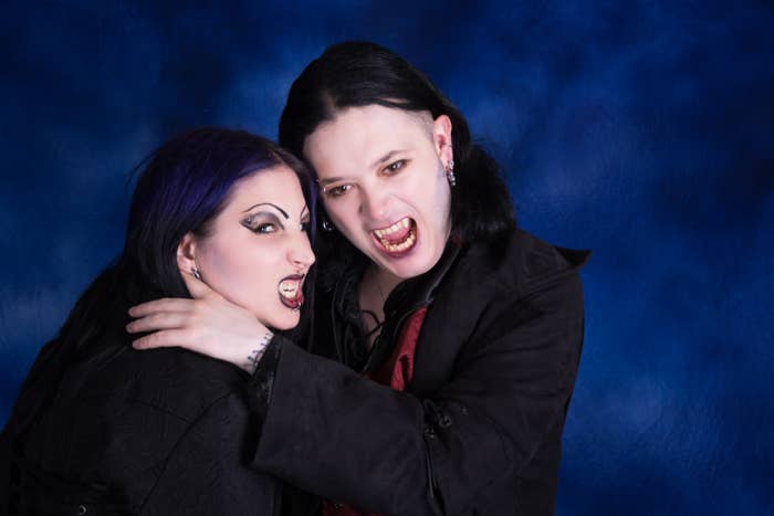 Two people with vampire-like makeup and fangs pose playfully against a blue backdrop, wearing dark gothic clothing