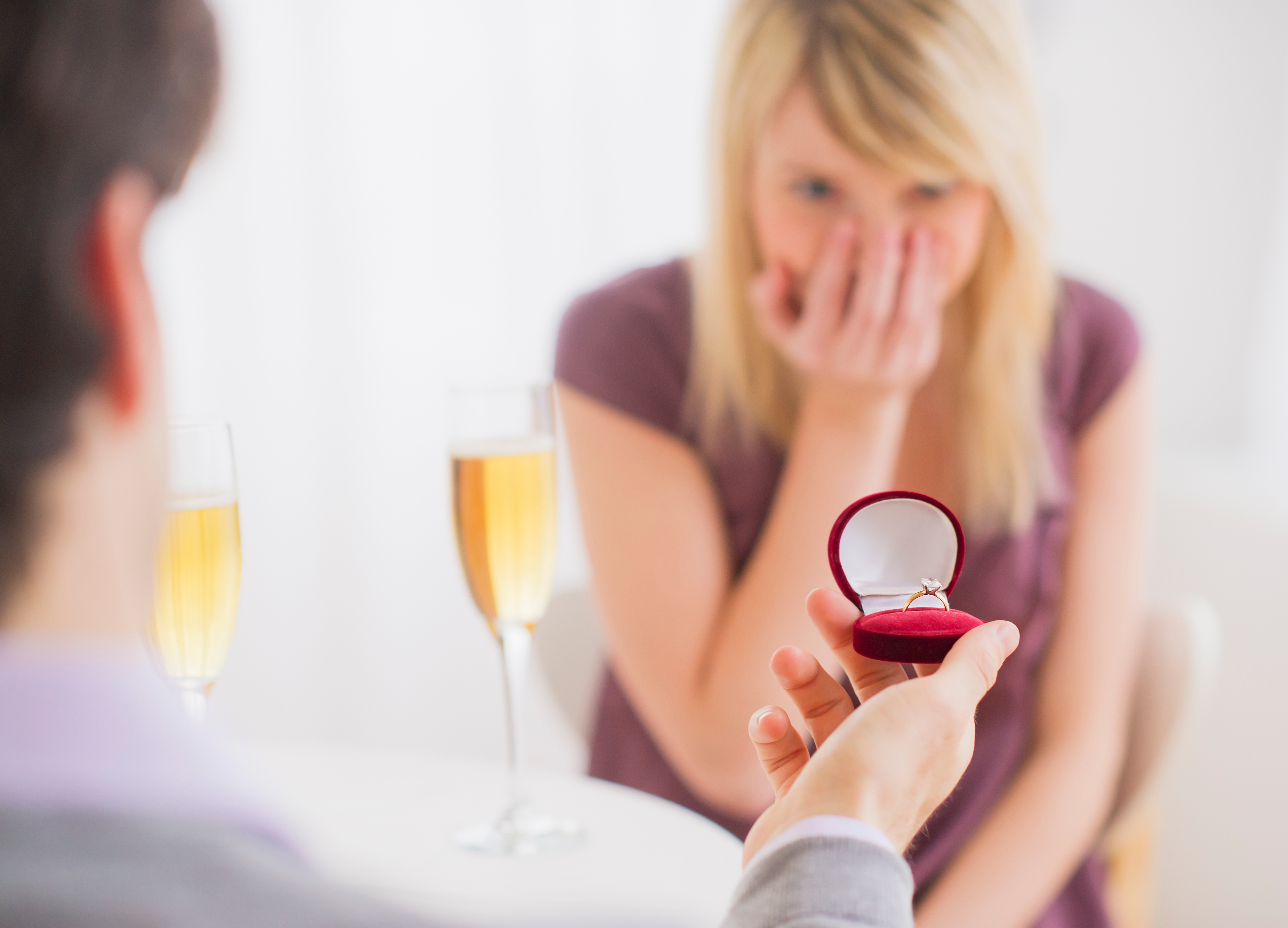 Person holds an open ring box while another person looks surprised, with two glasses of champagne on the table