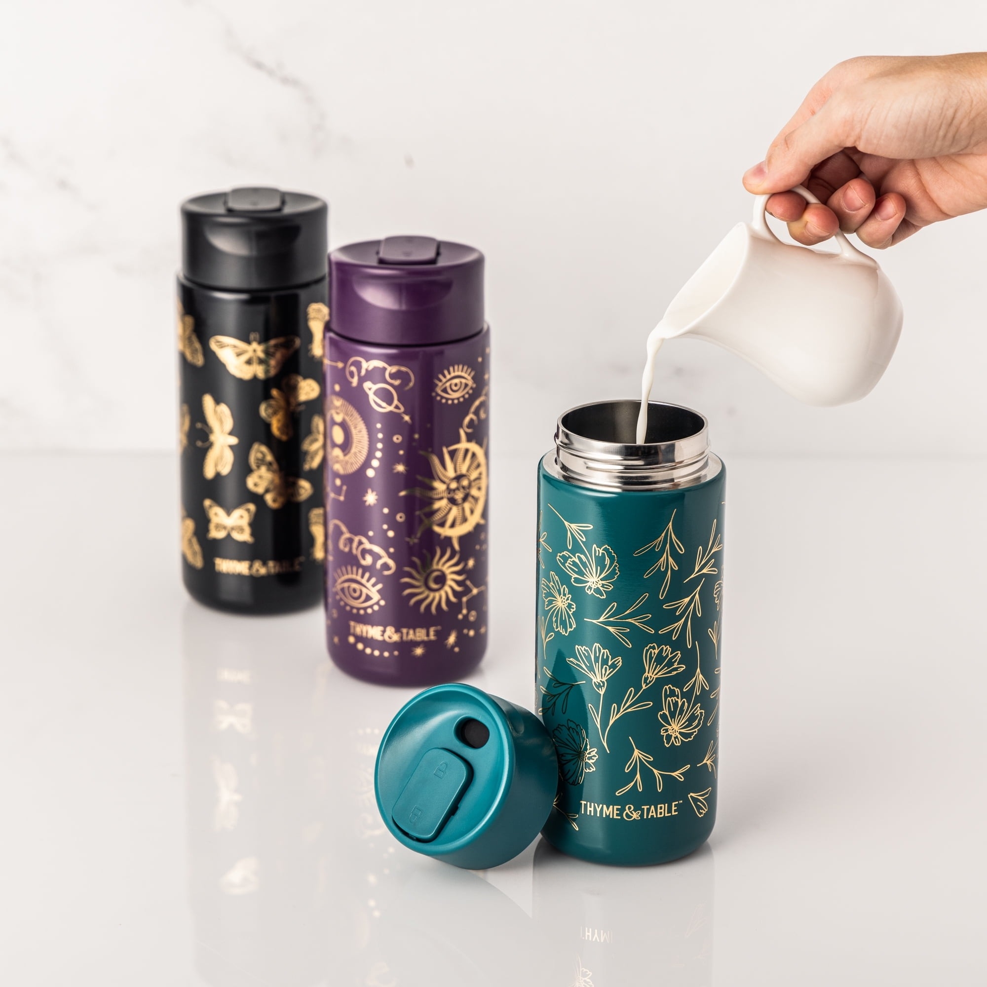 Three decorative insulated bottles in black, purple and teal with gold designs; a hand pours cream into the teal bottle with floral design