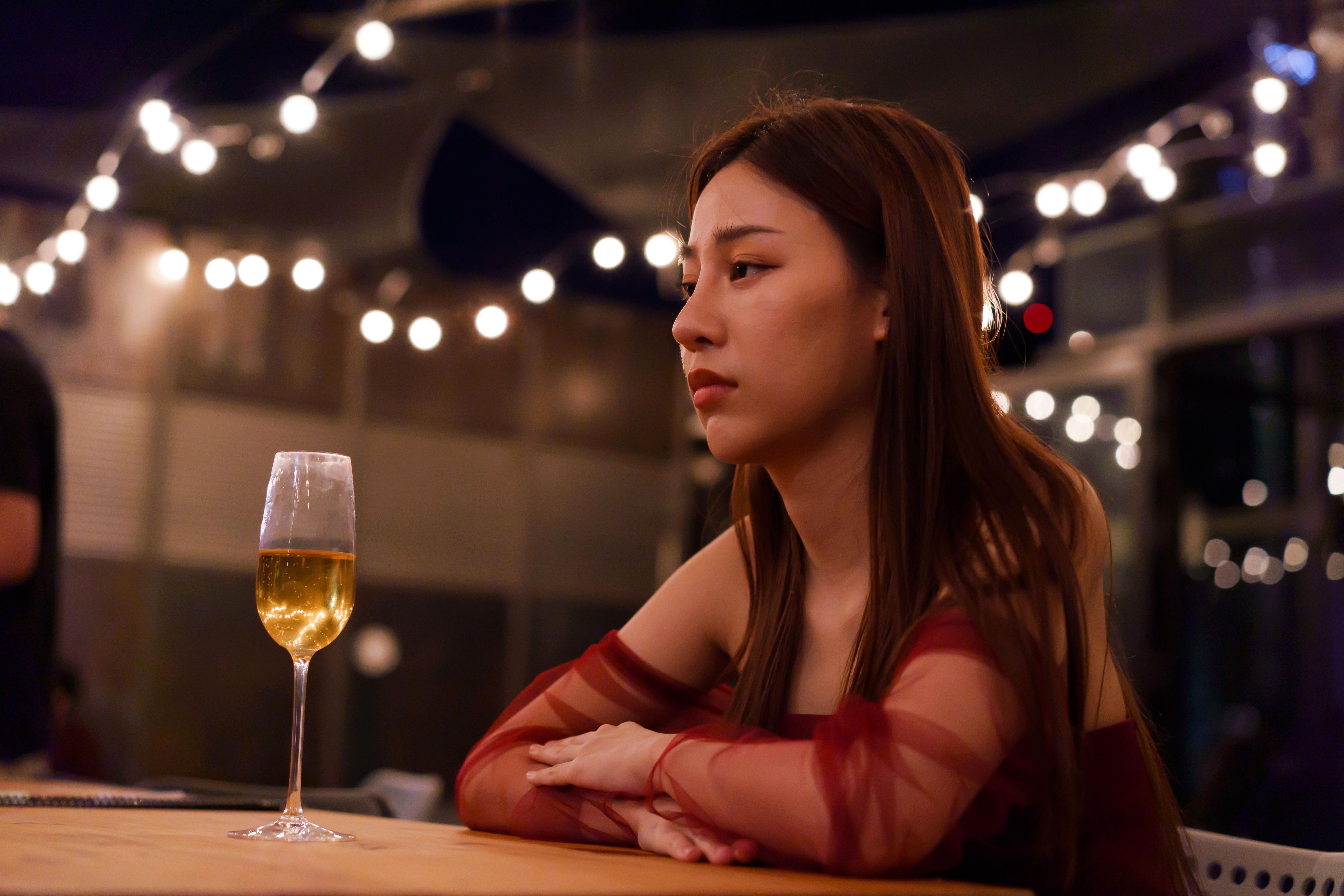A person sits at a bar with string lights in the background, looking contemplative. A glass of champagne is on the table