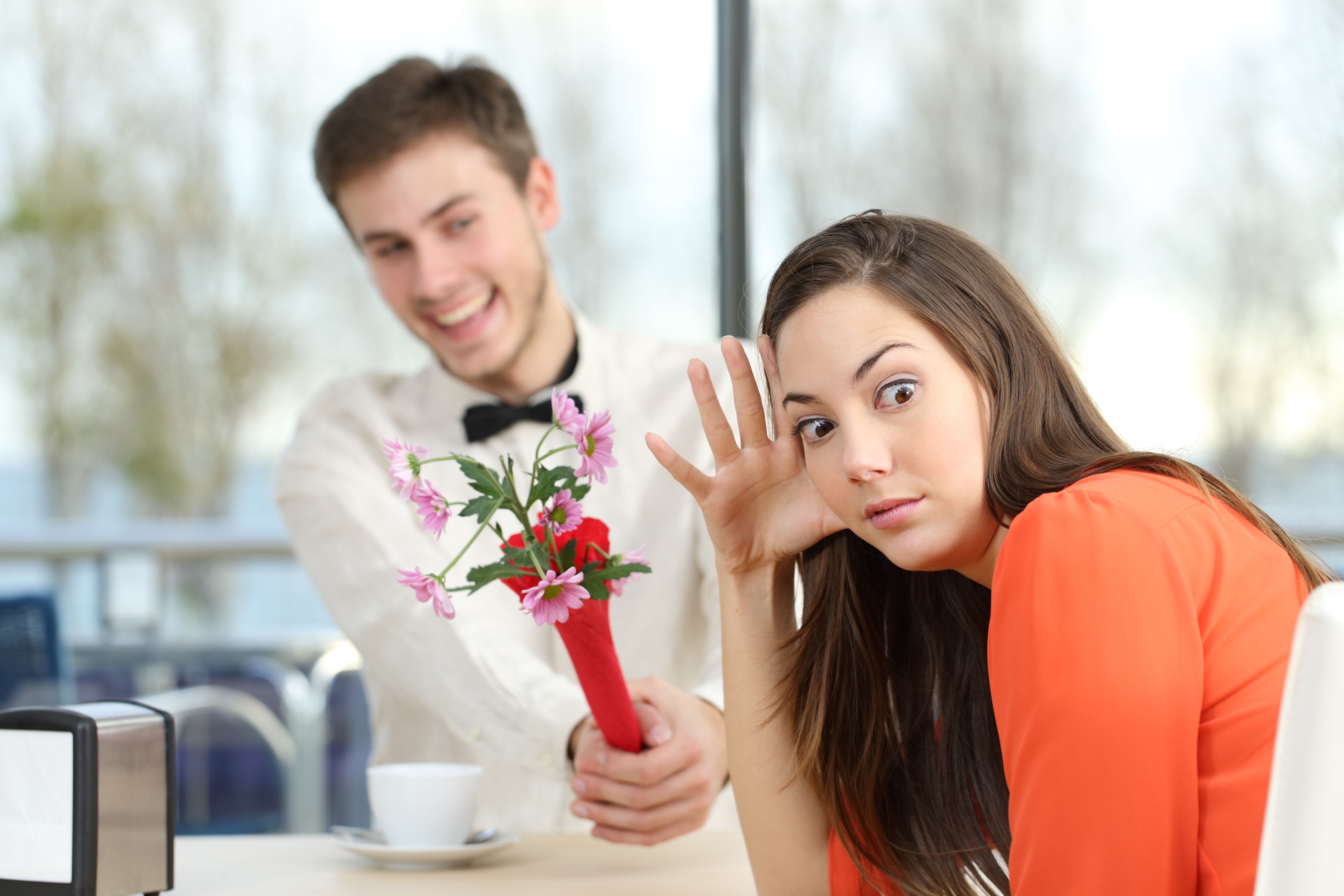 Man playfully offering flowers to an uninterested woman in a café setting, symbolizing unrequited love