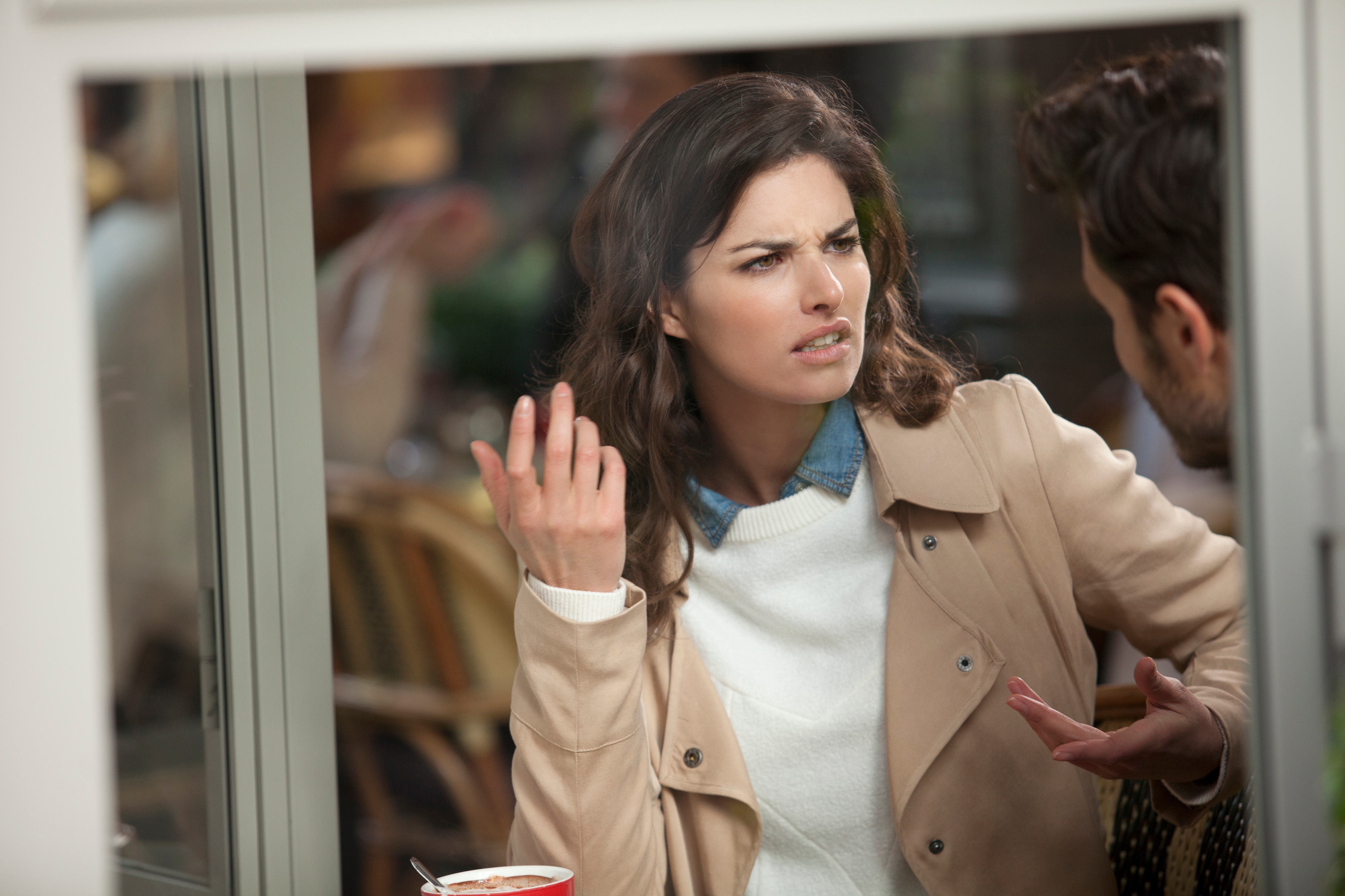 A woman in a cafe looks frustrated, gesturing while talking to a man seated across from her, suggesting a tense conversation