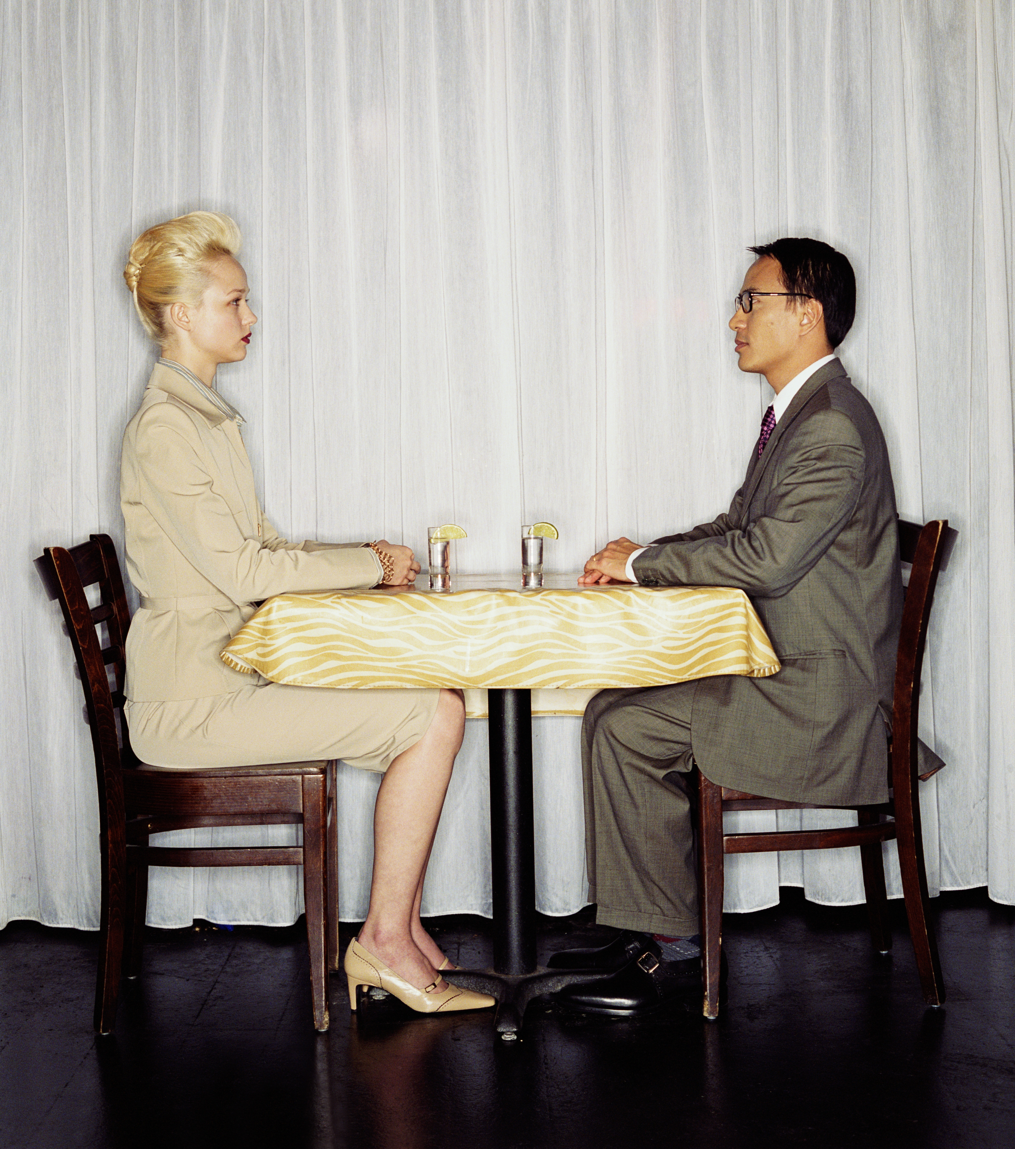 Two people in business attire sit across a small table with two drinks, facing each other in a poised and formal manner
