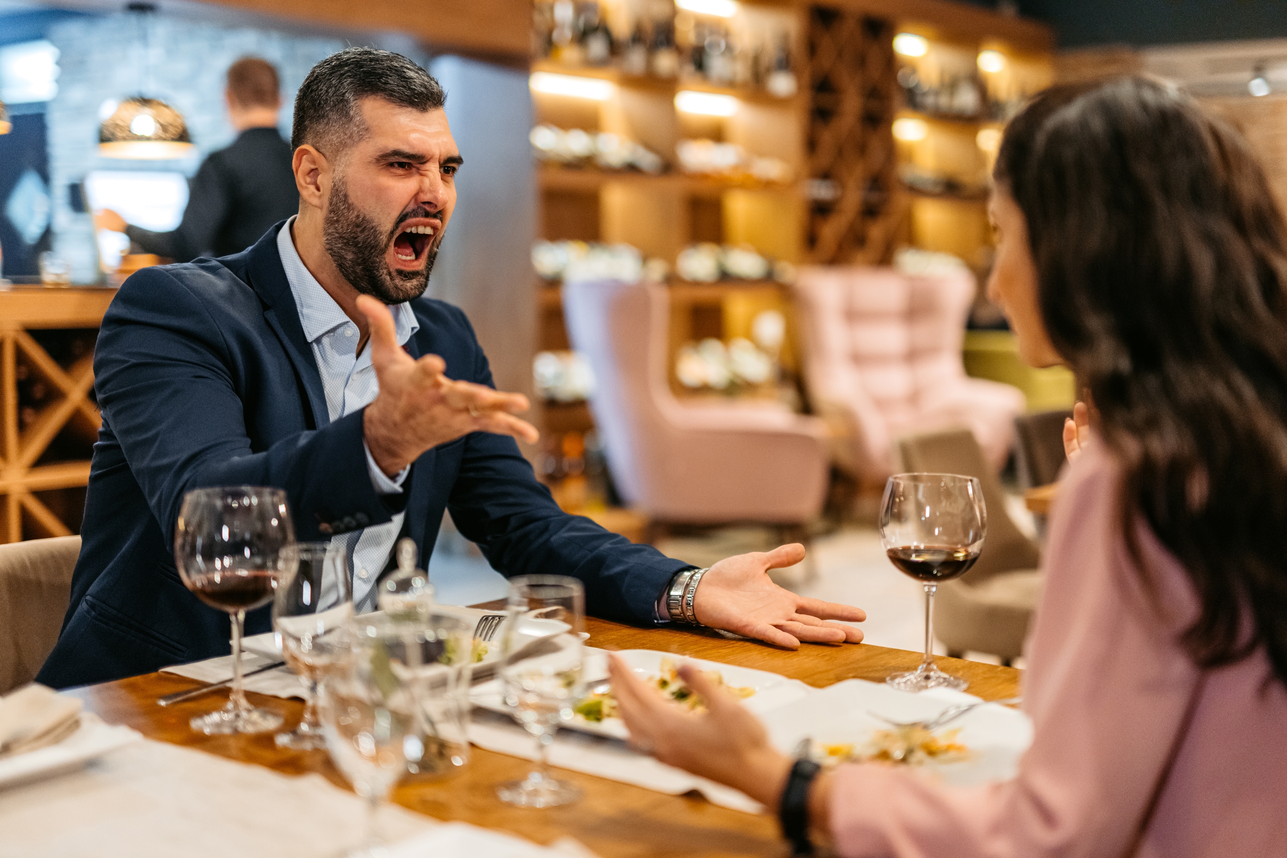 A man in a suit angrily gestures during a heated dinner conversation with a woman in a restaurant