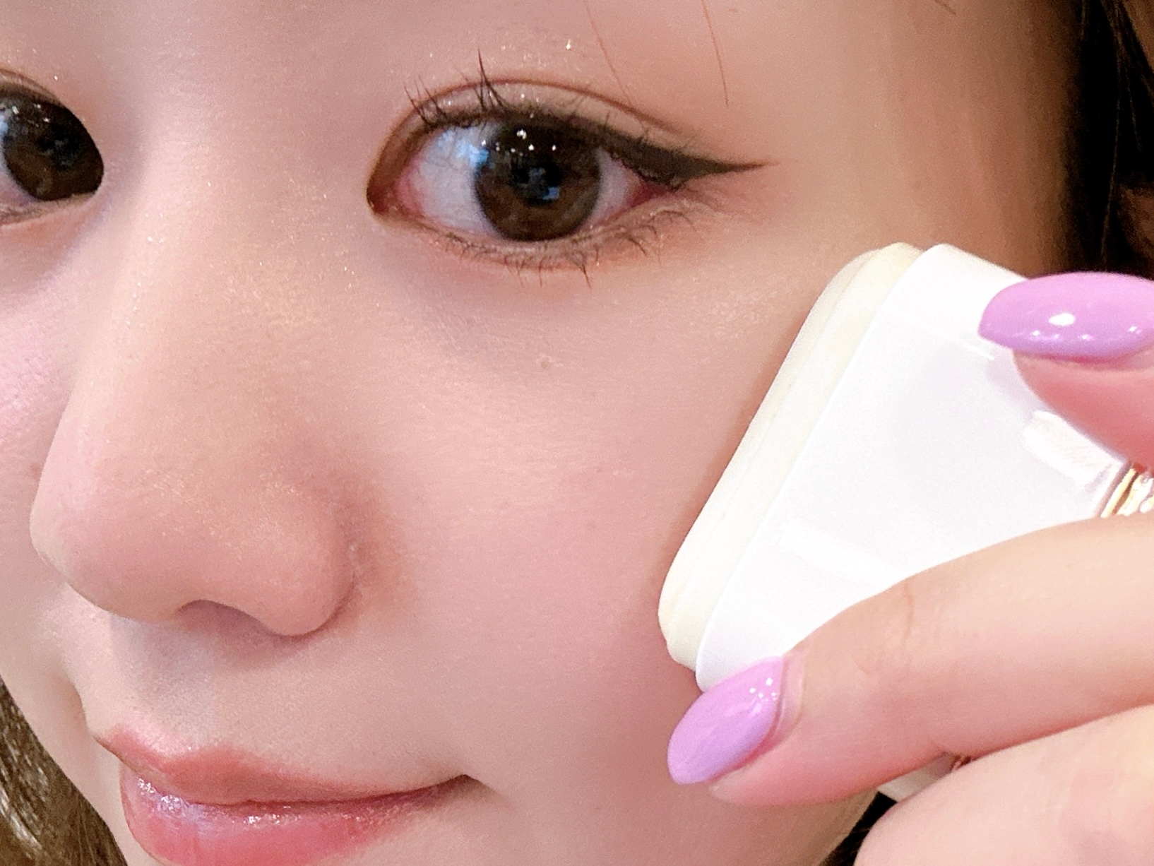 女性が顔に化粧パフを当てているクローズアップ写真。指のネイルは紫色。