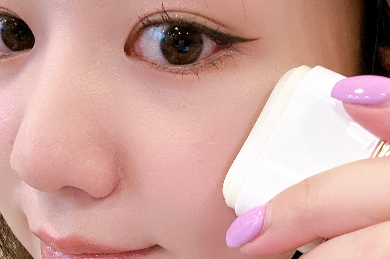 女性が顔に化粧パフを当てているクローズアップ写真。指のネイルは紫色。