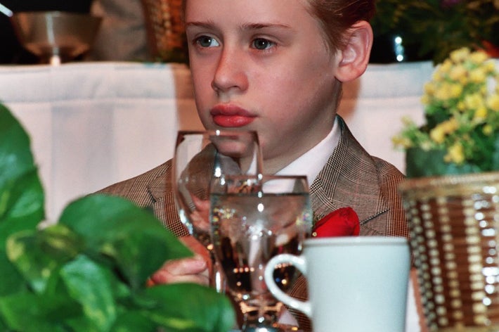 A young person in a formal suit sits at a table with plants and tableware, appearing thoughtful during an event